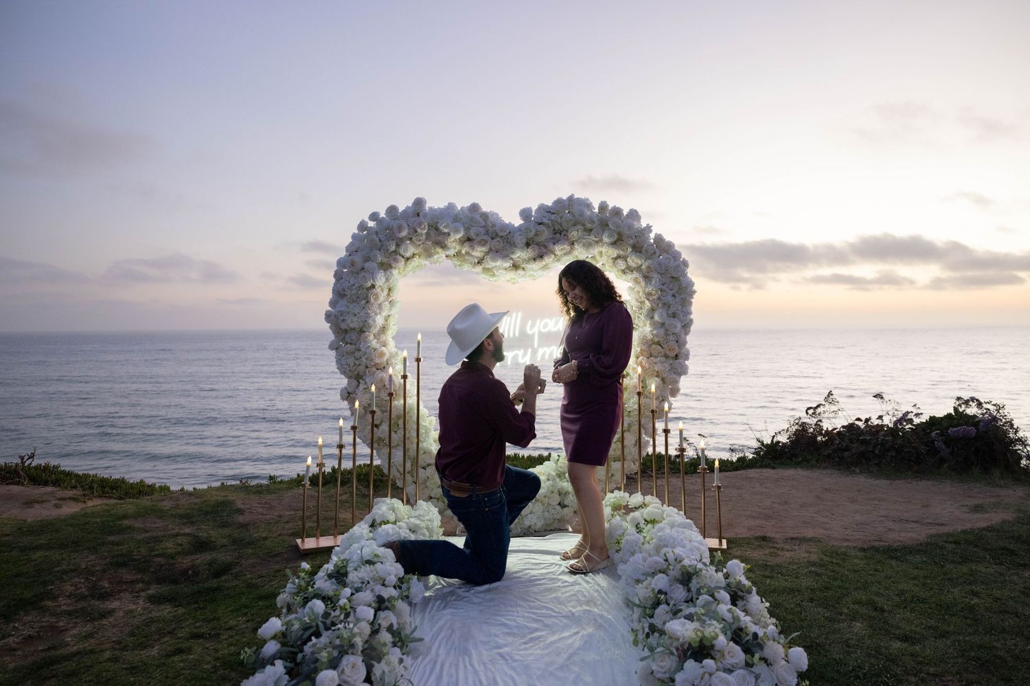 Romantic sunset proposal with heart-shaped floral arch overlooking the ocean, man on one knee presenting ring to woman. Engagement Photos.
