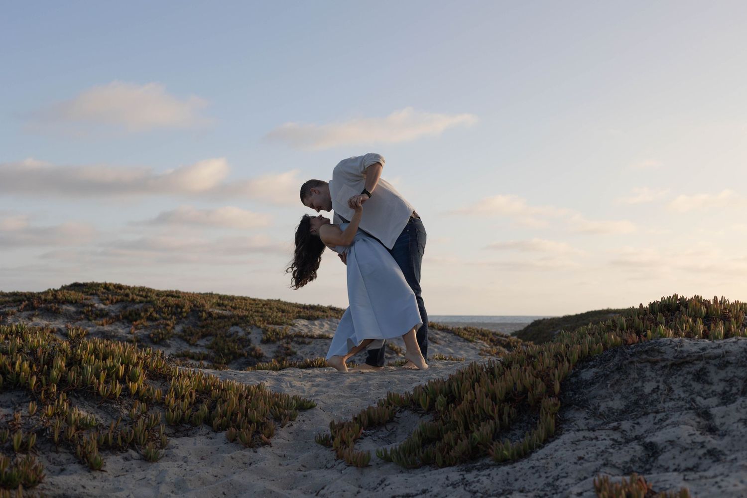 Couple sharing a romantic dip kiss on sandy dunes at sunset, surrounded by coastal vegetation under a pastel sky. Coronado Beach San Diego.