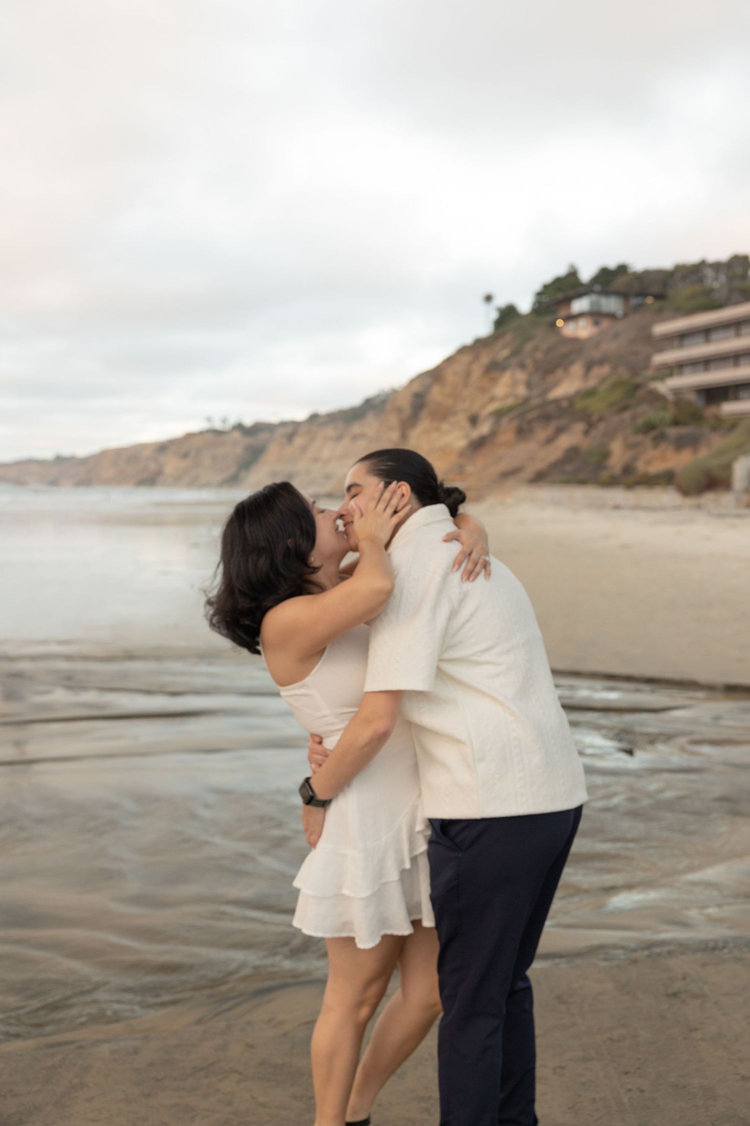 Couple sharing a romantic kiss on a sandy beach with coastal cliffs in the background.