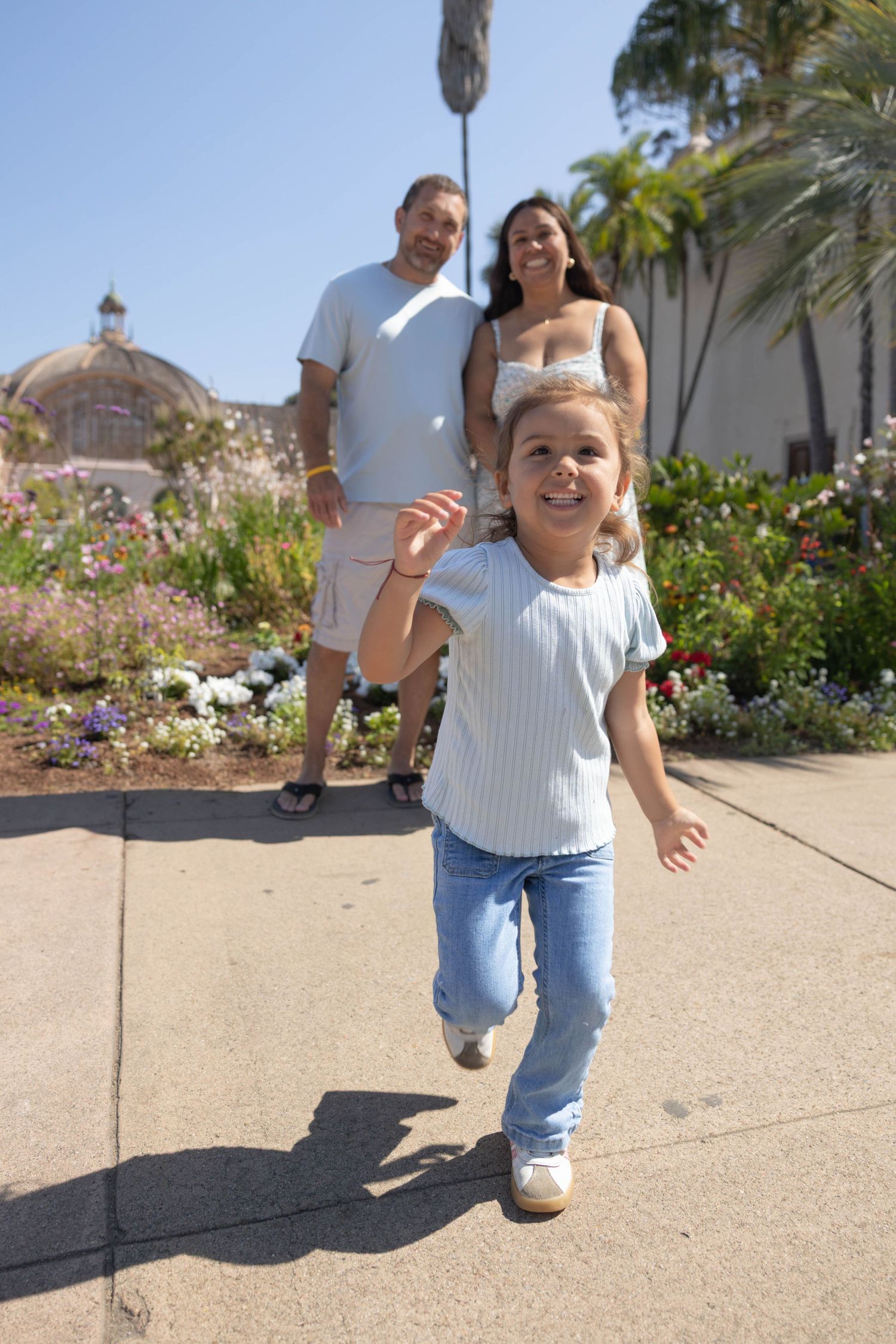 A young girl runs ahead of her smiling parents along a sunny garden path with colorful flowers and palm trees.