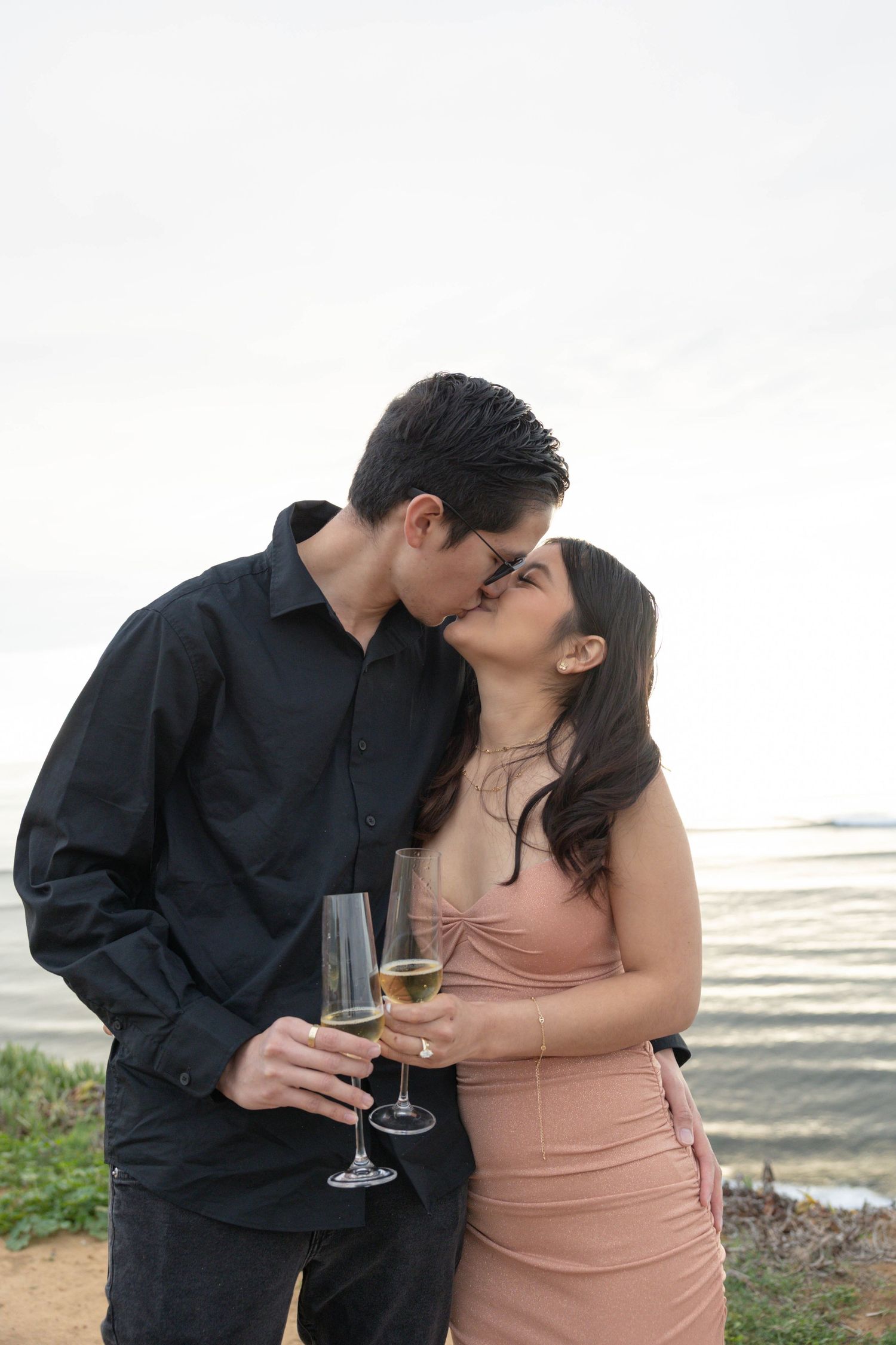 Couple kissing while holding champagne glasses near waterfront, man in black shirt, woman in pink dress. San Diego Engagement Photographer.