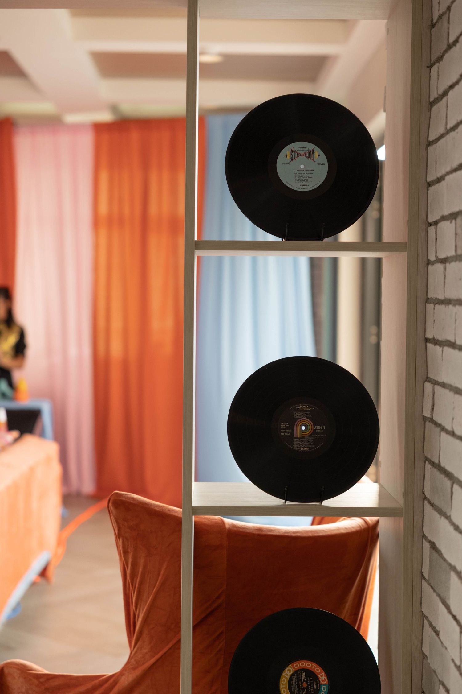 Two vintage vinyl records displayed on white shelf unit beside orange velvet chair and curtained window background.