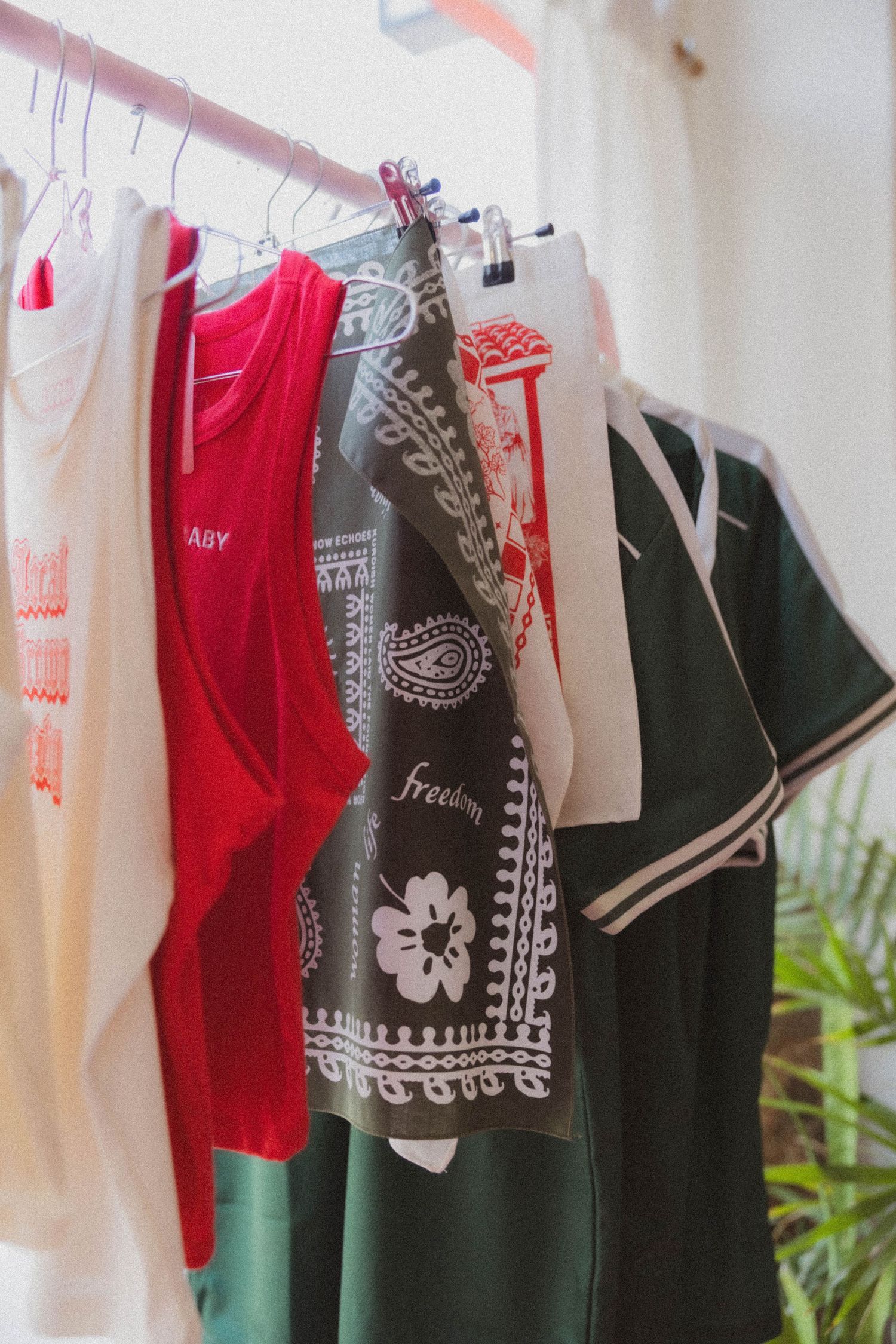 Clothing rack displaying red tank top, dark bandana print apron, and green varsity jacket in bright boutique.