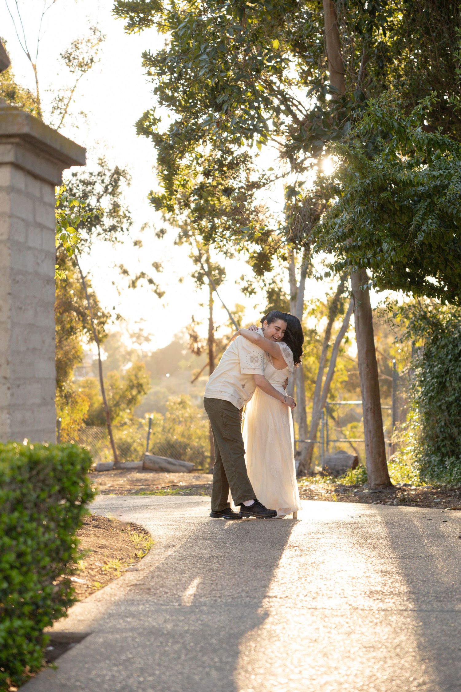 Couple embracing on a sunlit garden path surrounded by lush trees in warm golden hour light.