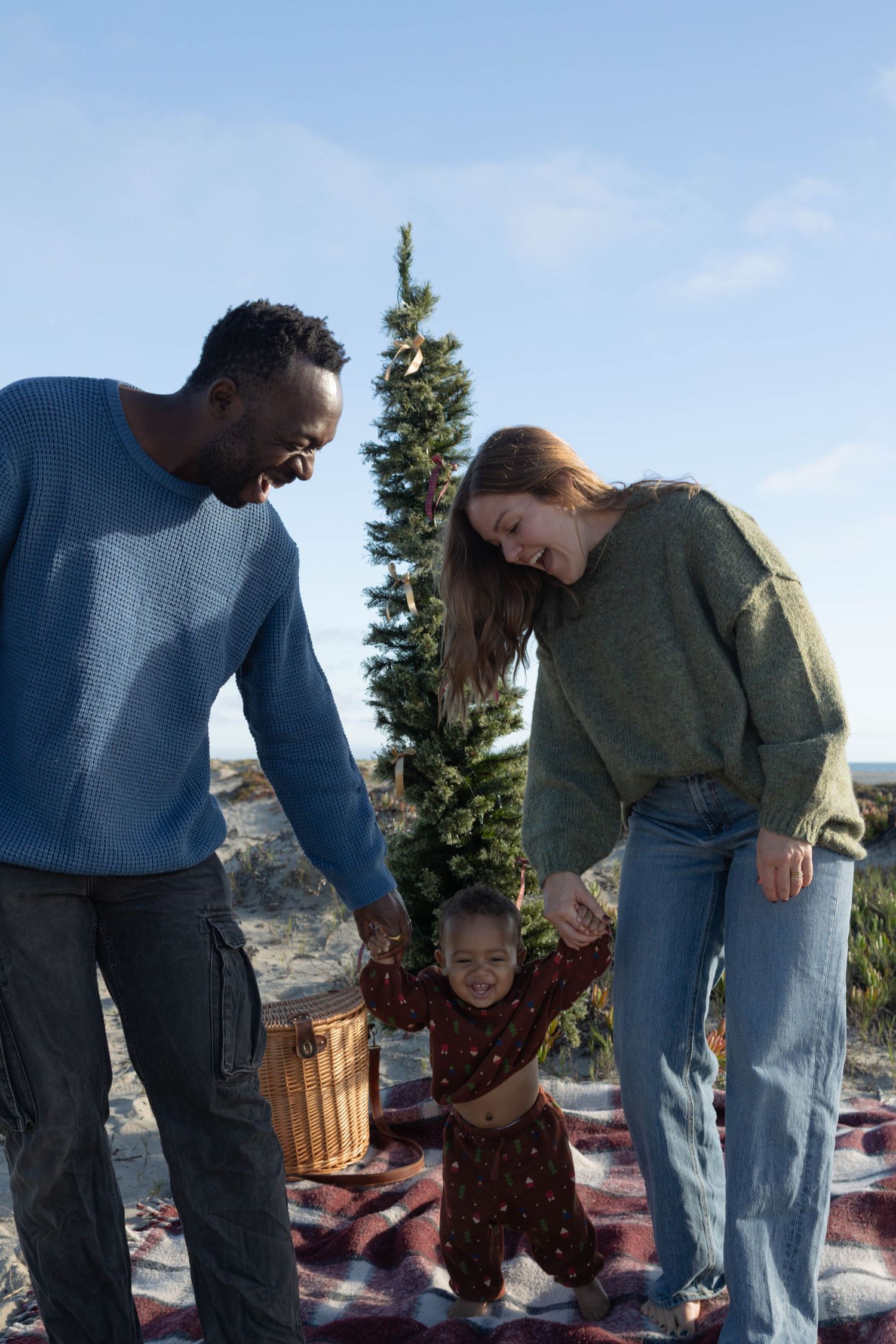 Interracial family with toddler holding hands outdoors near Christmas tree on blanket, smiling joyfully.