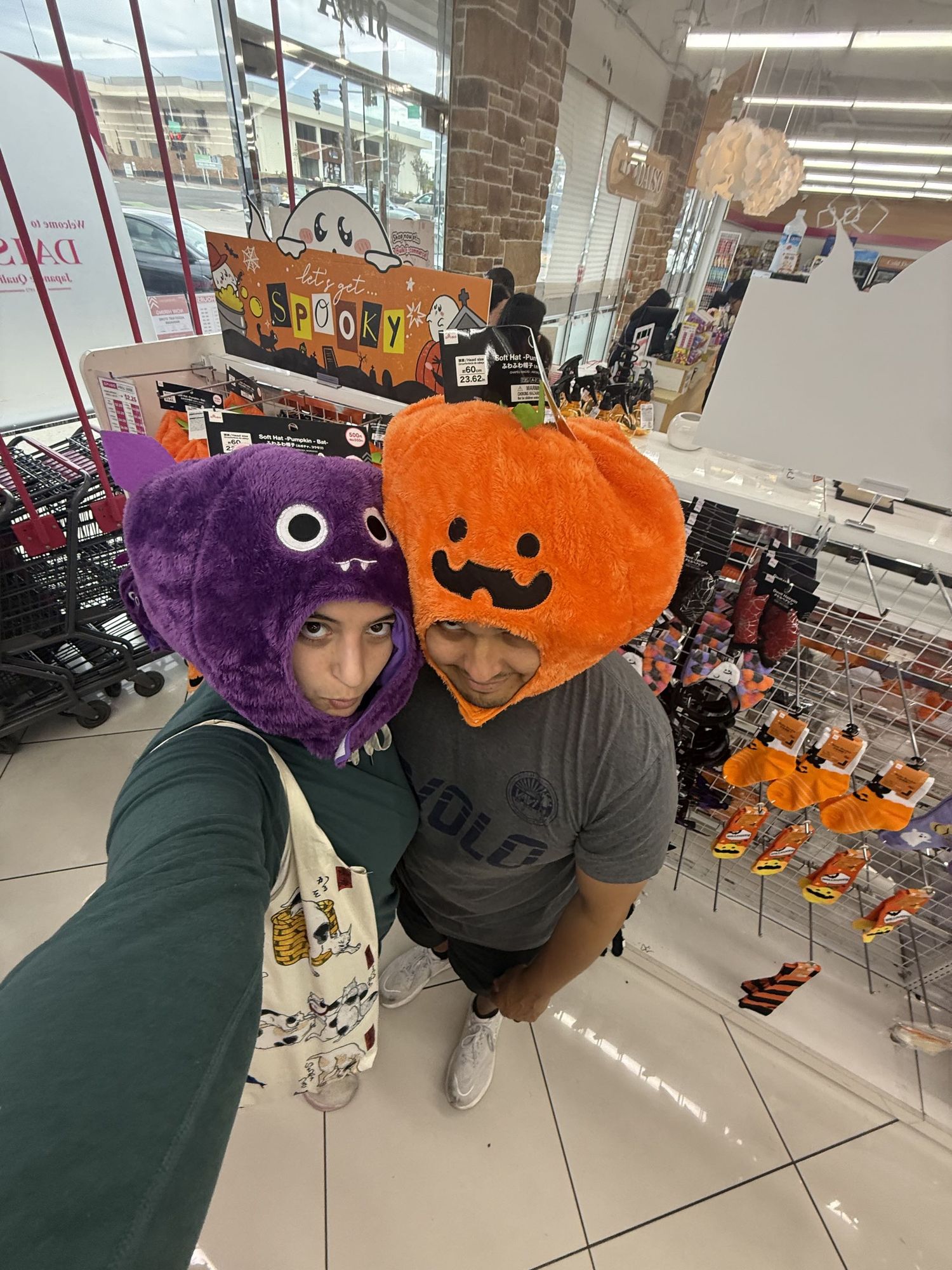 Two people wearing Halloween hats, a purple monster and orange pumpkin, posing for a selfie in a festive store.