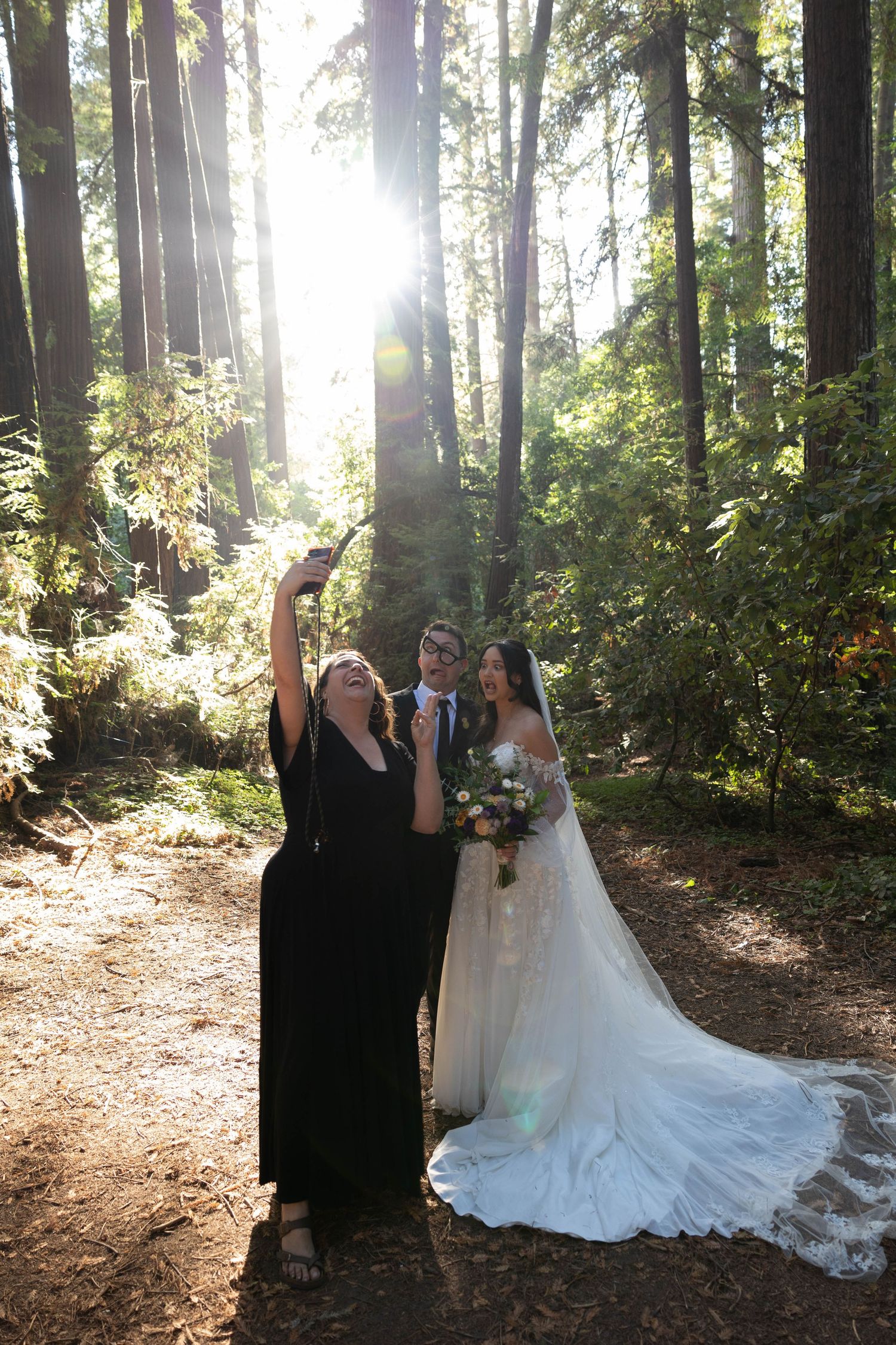 Bride and groom take a selfie with a guest in a sunlit redwood forest on their wedding day. Wedding photography.