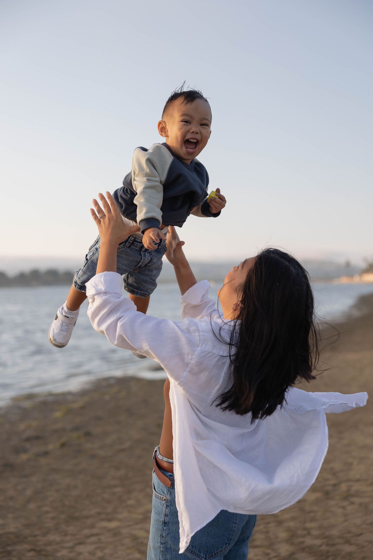 A joyful mother lifts her laughing baby high in the air on a sunny beach near the water. Family Photographer.