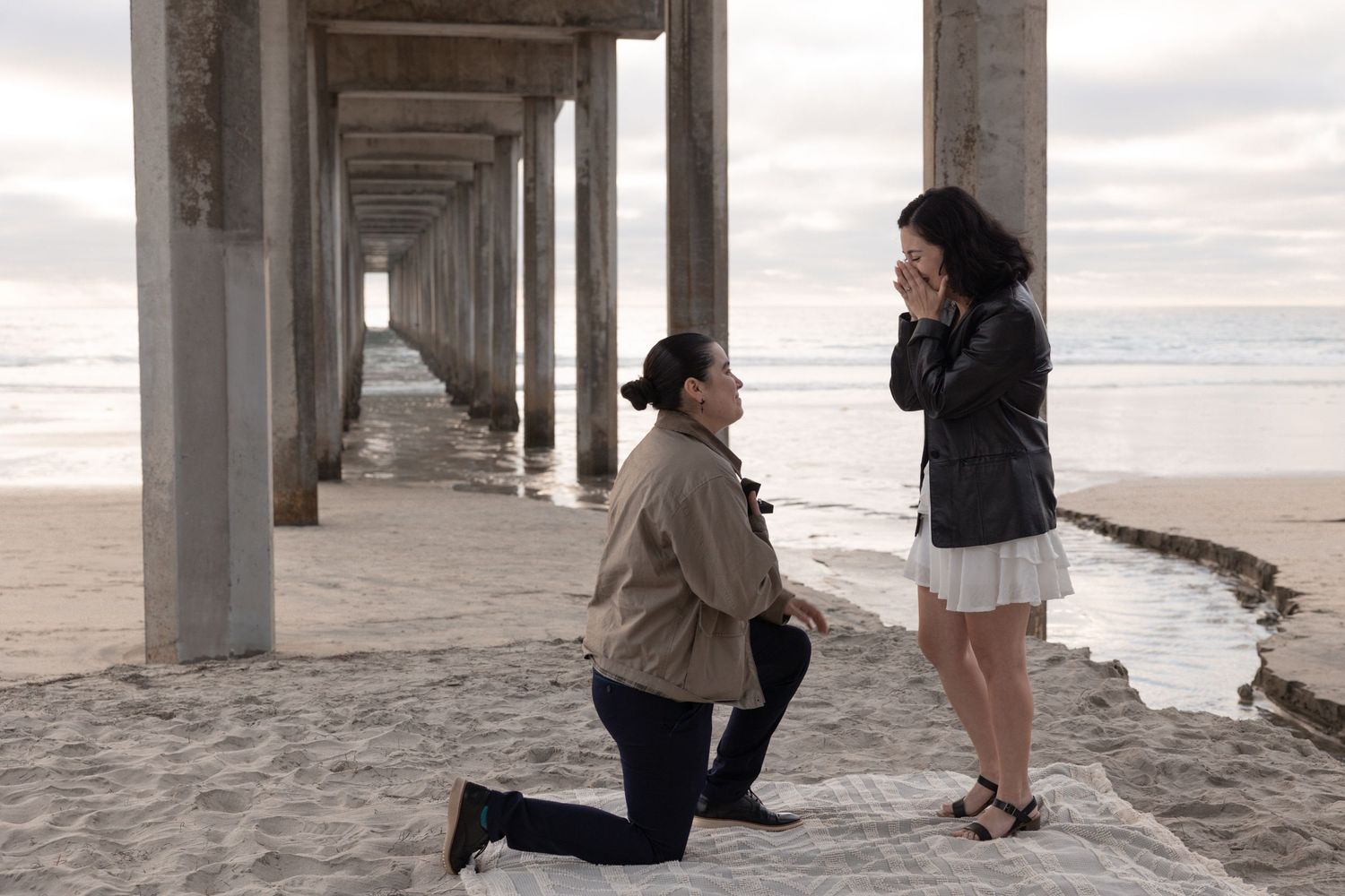 Person proposing on one knee to partner under a beach pier, with ocean waves in the background at dusk. San Diego Engagement photographer.