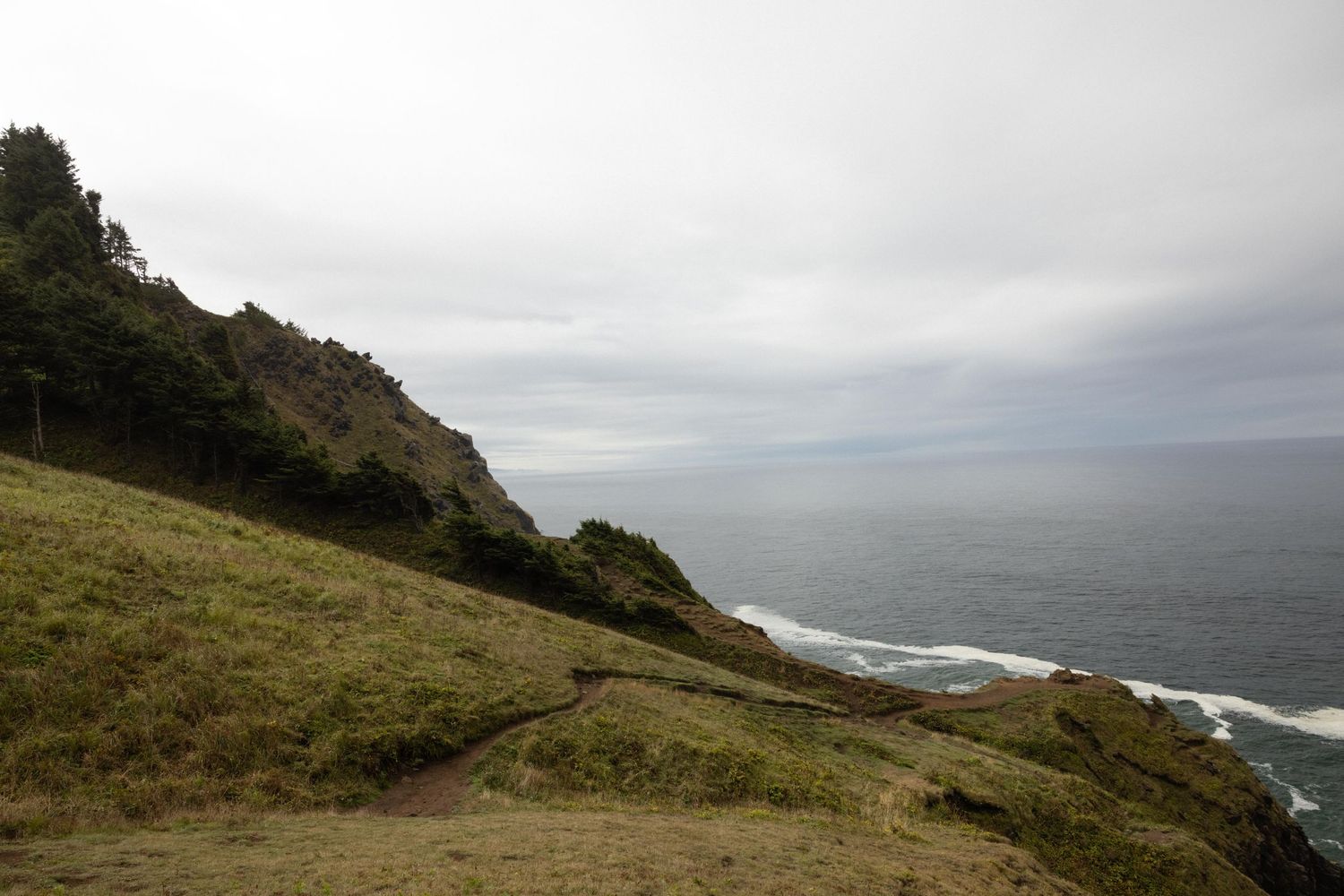 Dramatic coastal cliffside with lush green hills overlooking a vast ocean under overcast skies.