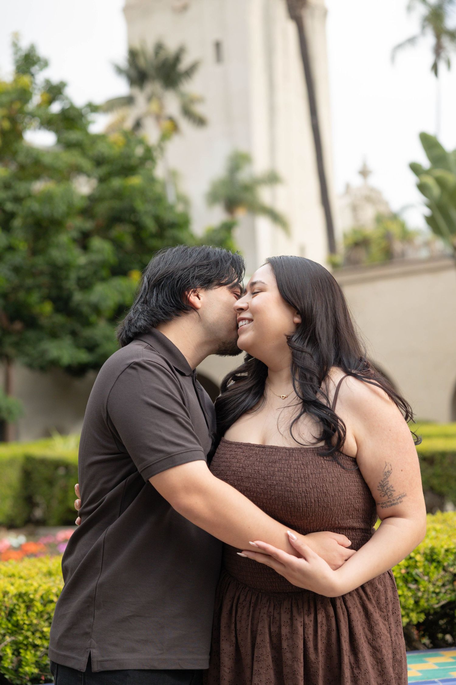 A couple sharing a romantic kiss in a garden setting with lush greenery and a white tower in the background. Couples Portraits San Diego Ca.