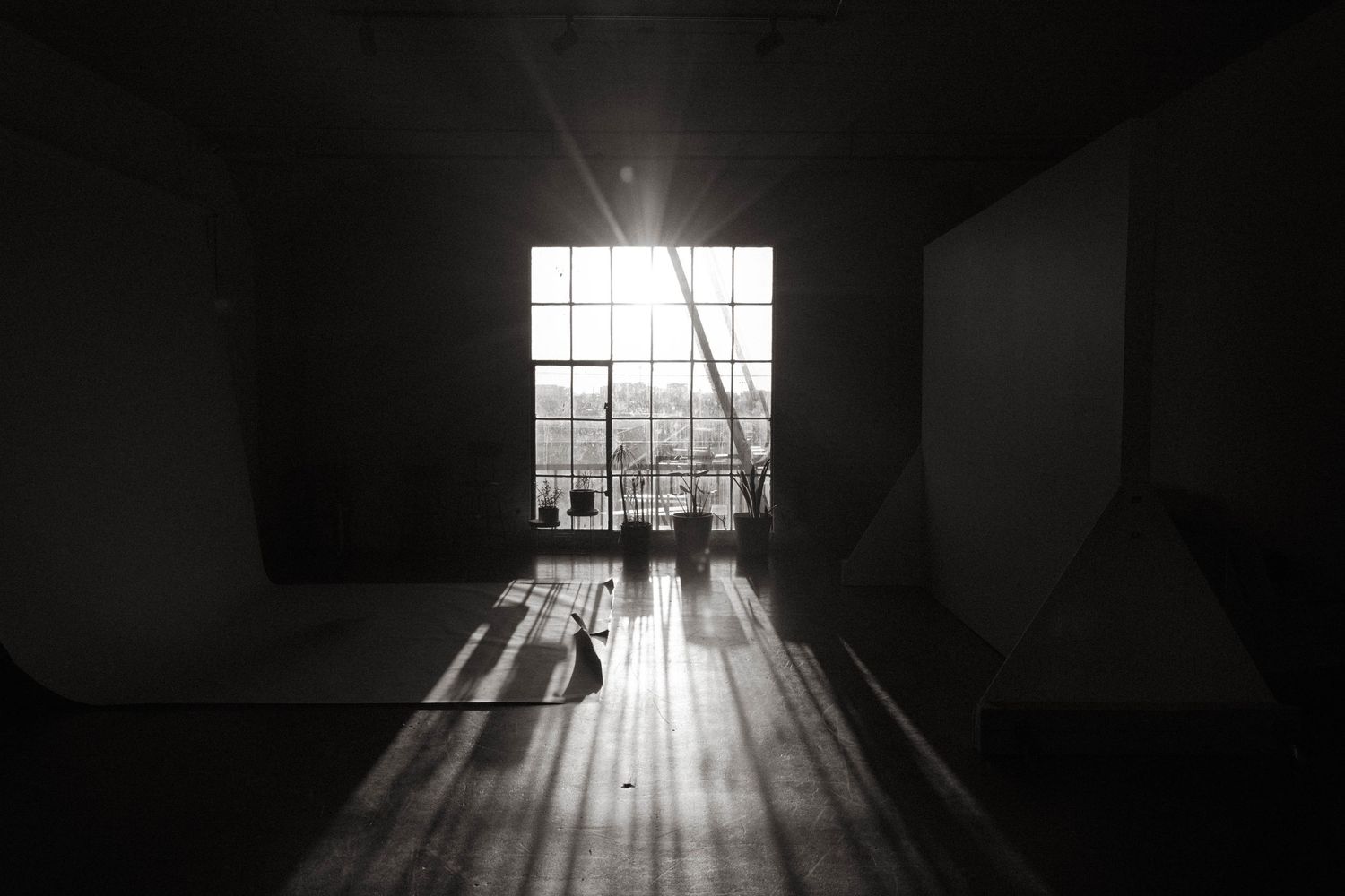 Black and white photo of a dark empty room with sunlight streaming through large industrial windows onto wooden floors.