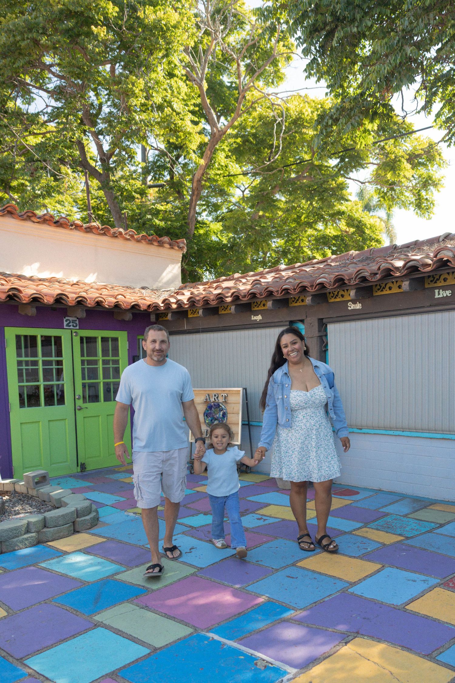 Family of three holding hands on colorful tiled courtyard with Spanish-style buildings and lush green trees.