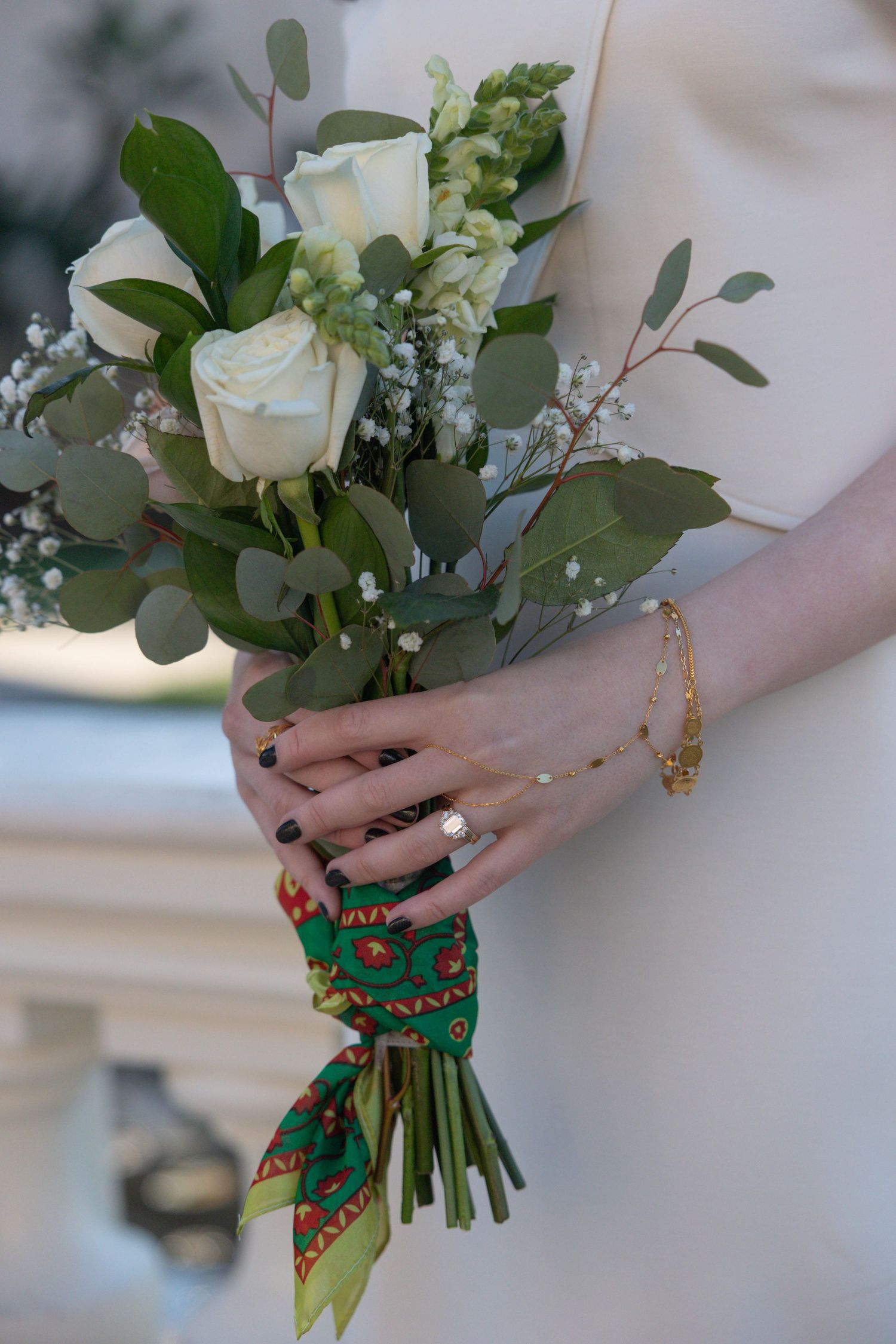 Close-up of bride holding white rose and eucalyptus bouquet with green patterned wrap, wearing diamond ring and gold bracelet.