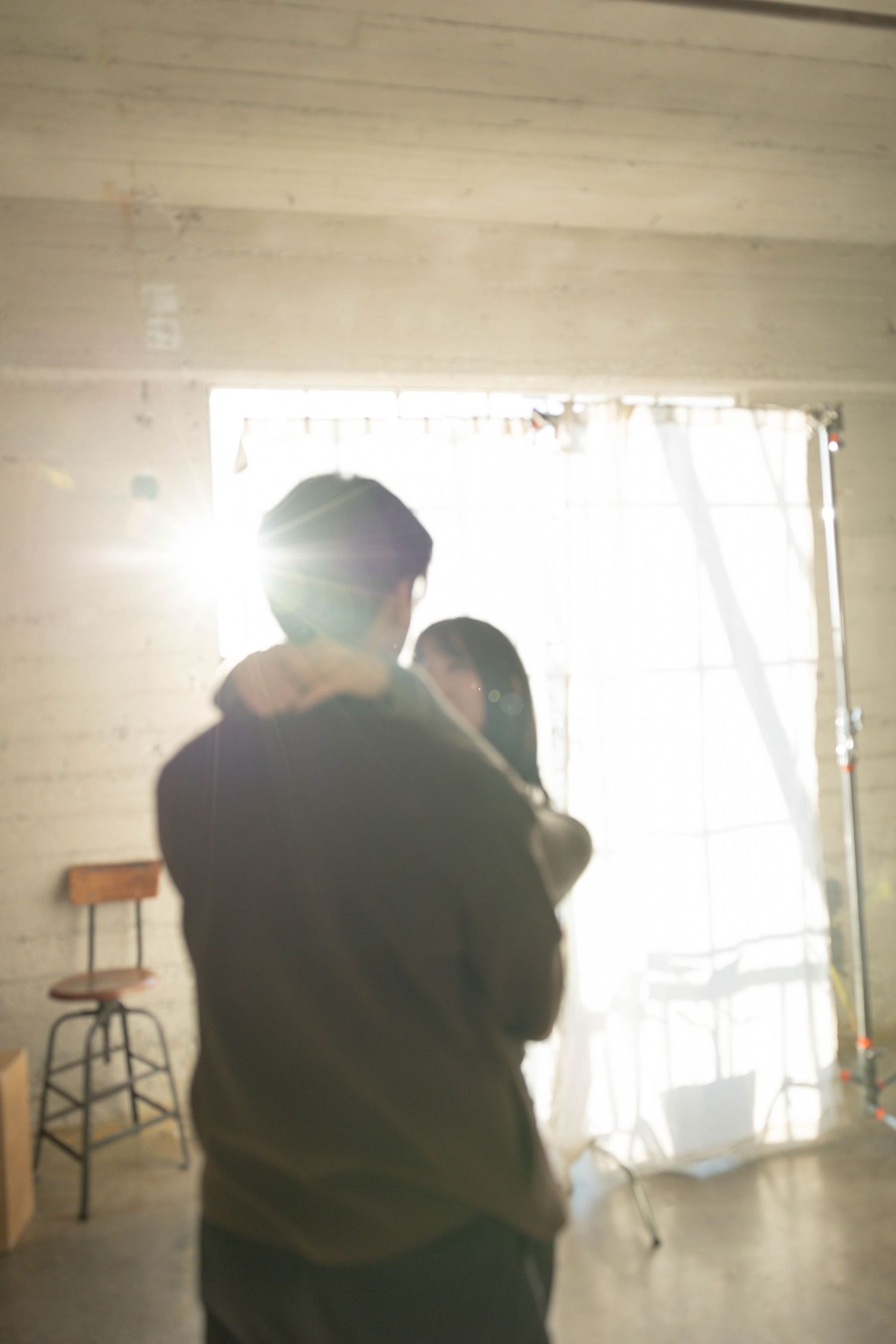 Couple embracing in silhouette against bright sunlit window in a warmly lit indoor studio space.