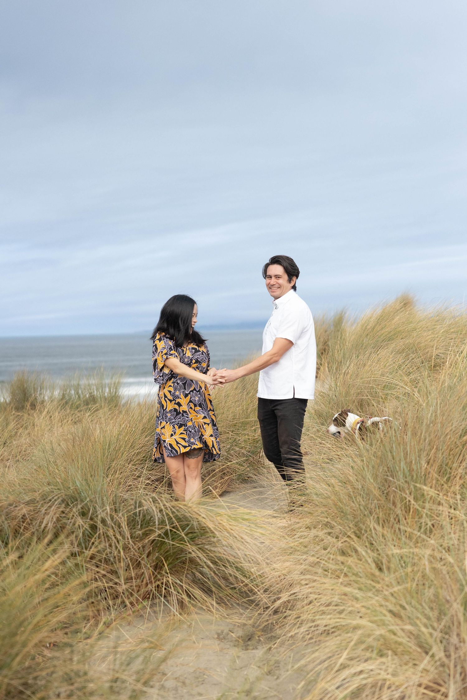 Couple holding hands on sandy beach dunes with tall grass and a dog, ocean in the background on a cloudy day. Maternity Photographer.
