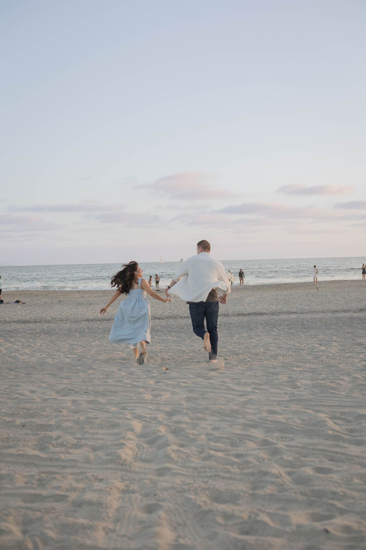 A couple runs hand-in-hand toward the ocean on a sandy beach at sunset, the woman in a light blue dress.