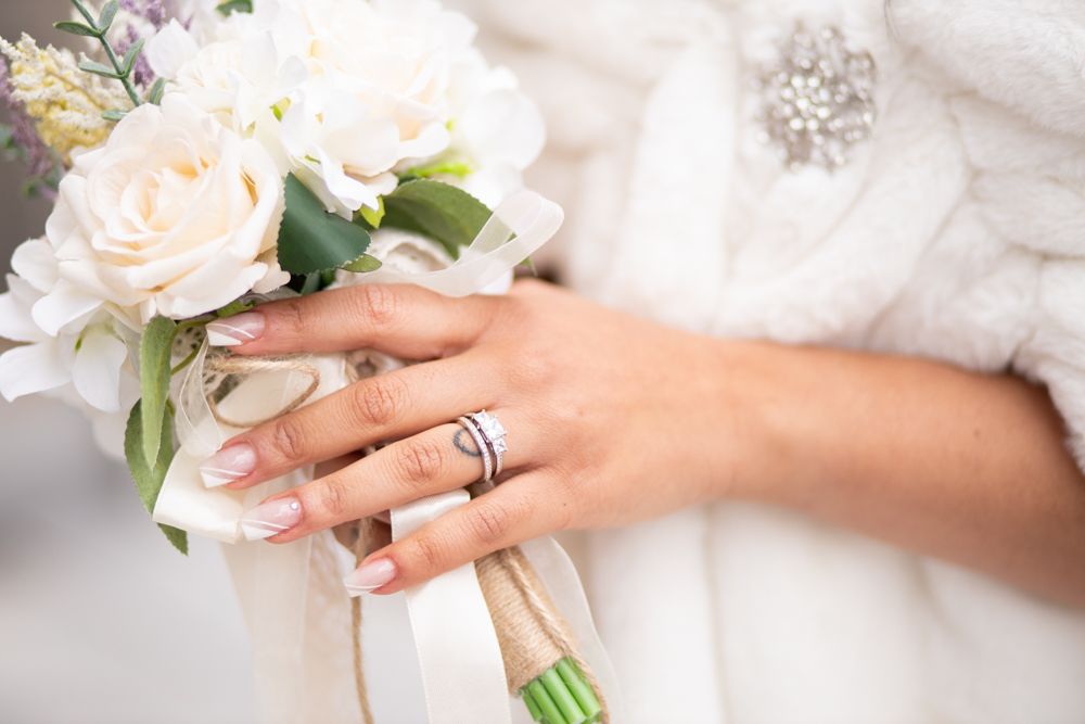 Hand wearing wedding rings holds elegant white rose bridal bouquet against white lace fabric background.