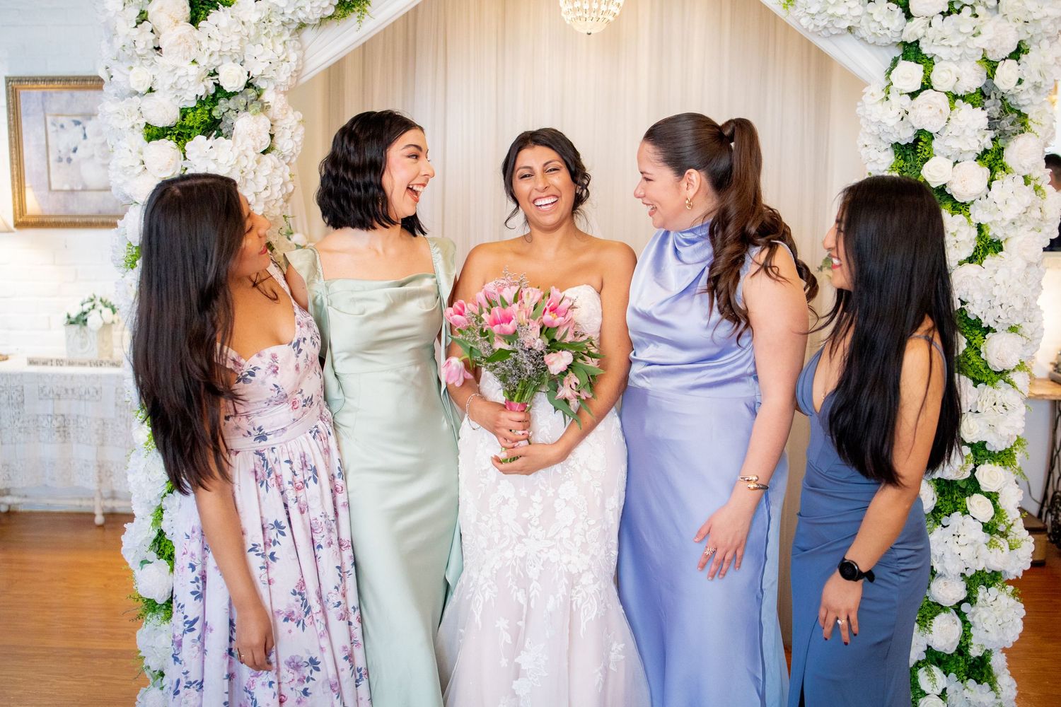 Bridesmaids in pastel purple and floral dresses pose together against a white floral arch backdrop at a wedding.