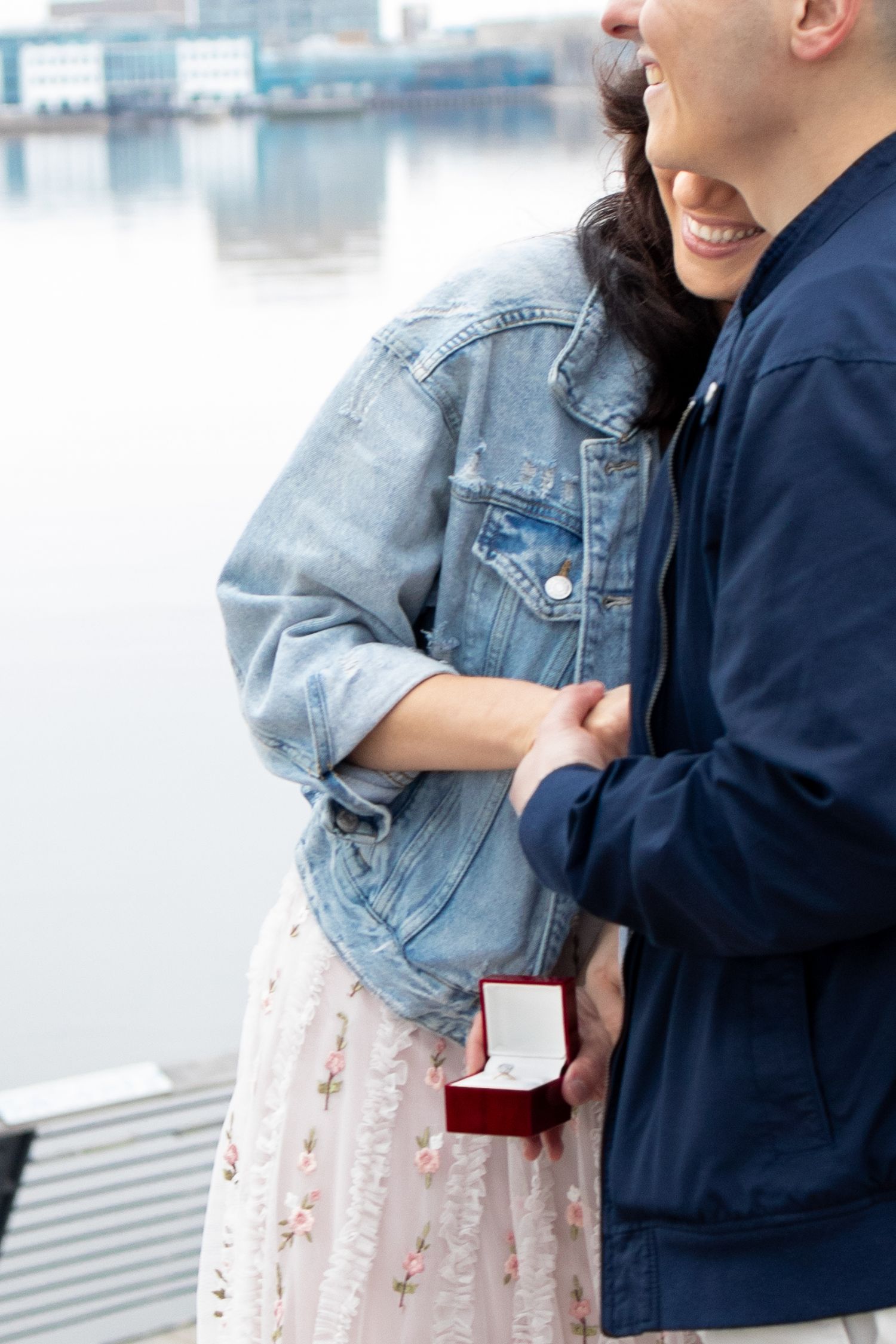 A romantic waterfront proposal moment with a denim jacket and floral dress, featuring a red ring box.