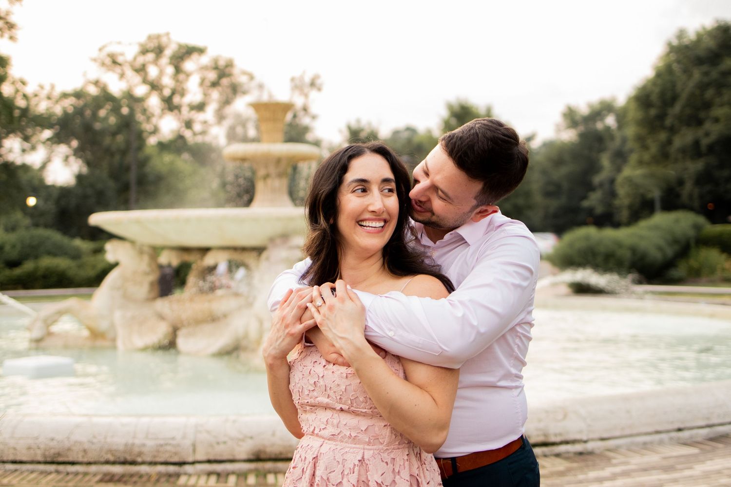 Couple embraces near ornate fountain in garden setting during sunset while wearing pink dress and white shirt.