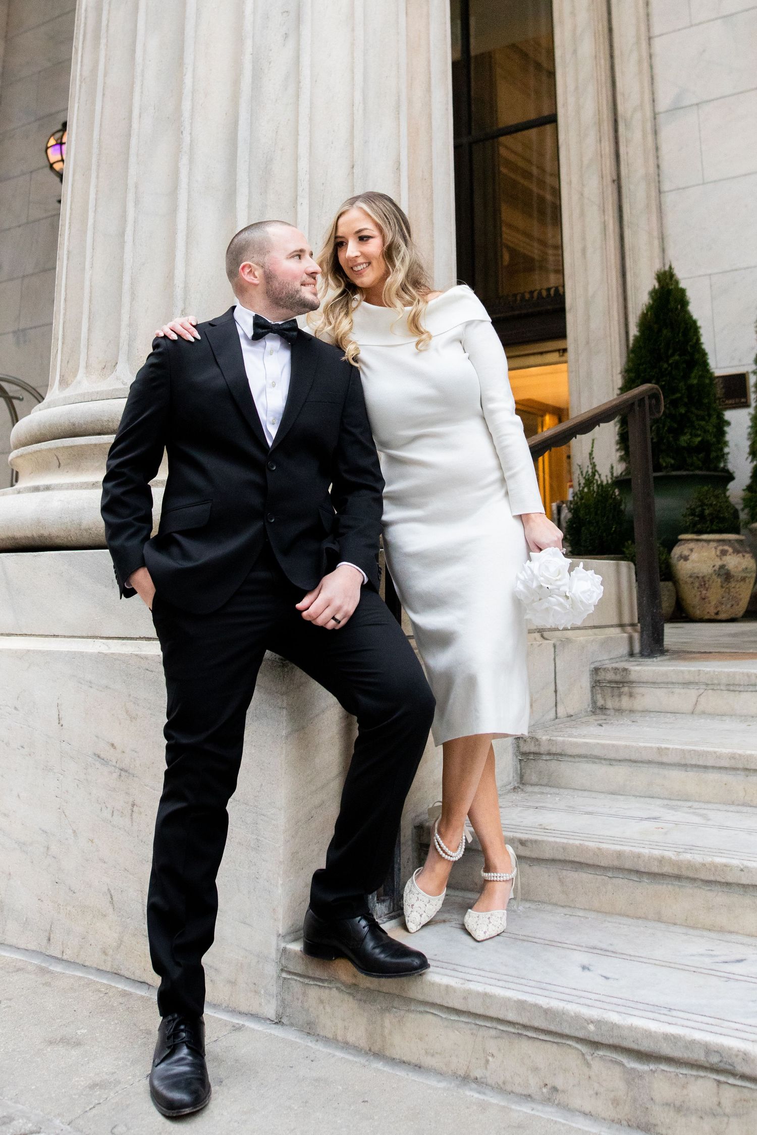 A couple dressed in elegant wedding attire pose together on marble steps outside a grand building.