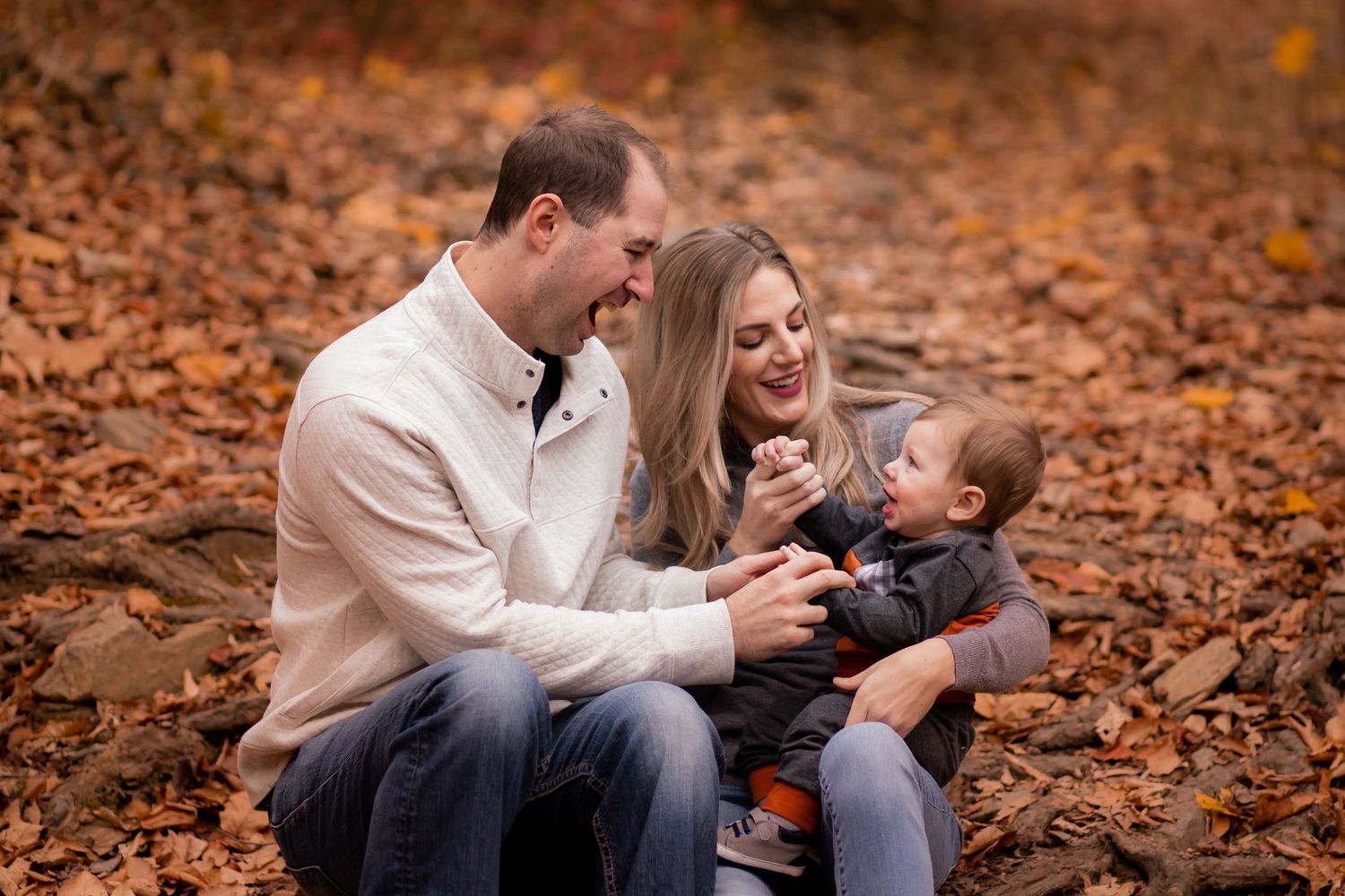 Family laughing and spending time together while sitting on fallen autumn leaves in a park.