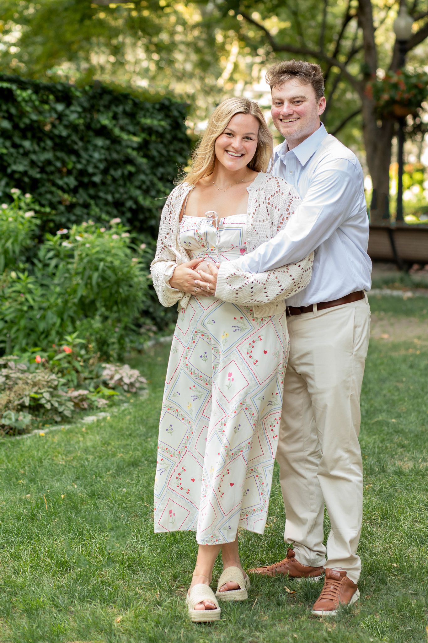 A couple poses together outdoors in a garden setting, wearing coordinated white and khaki summer outfits.