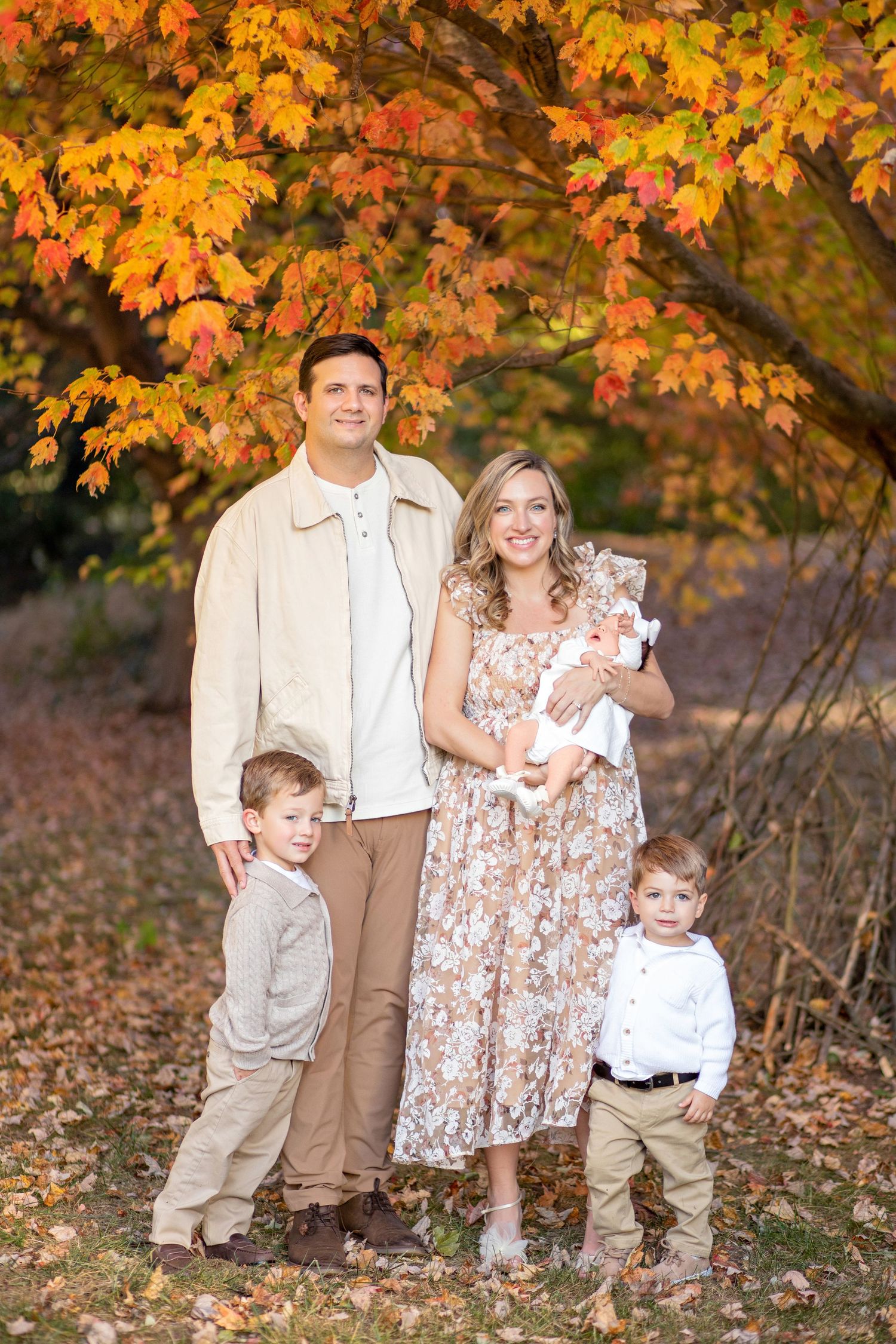 A family in coordinated fall outfits poses beneath vibrant orange autumn maple leaves on a crisp fall day.