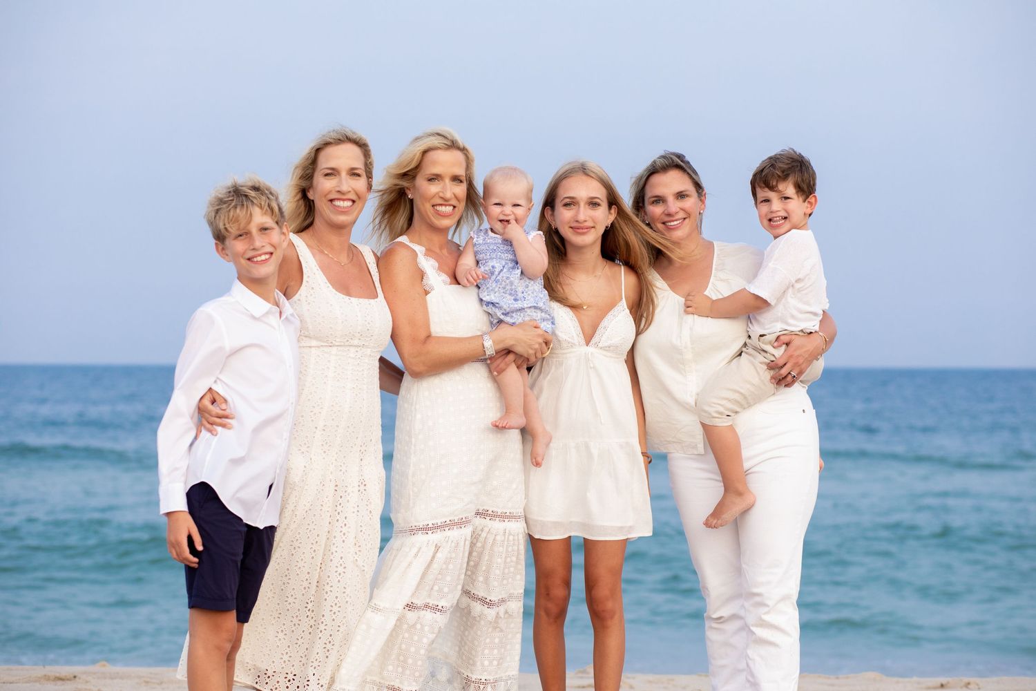 A group poses together on the beach wearing white clothing with the blue ocean in the background.