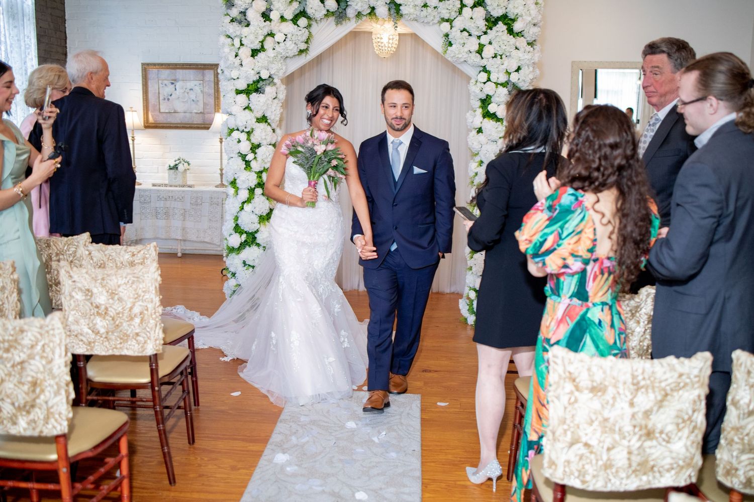 Wedding ceremony in a bright indoor venue with floral arch and elegant decor featuring guests in formal attire.