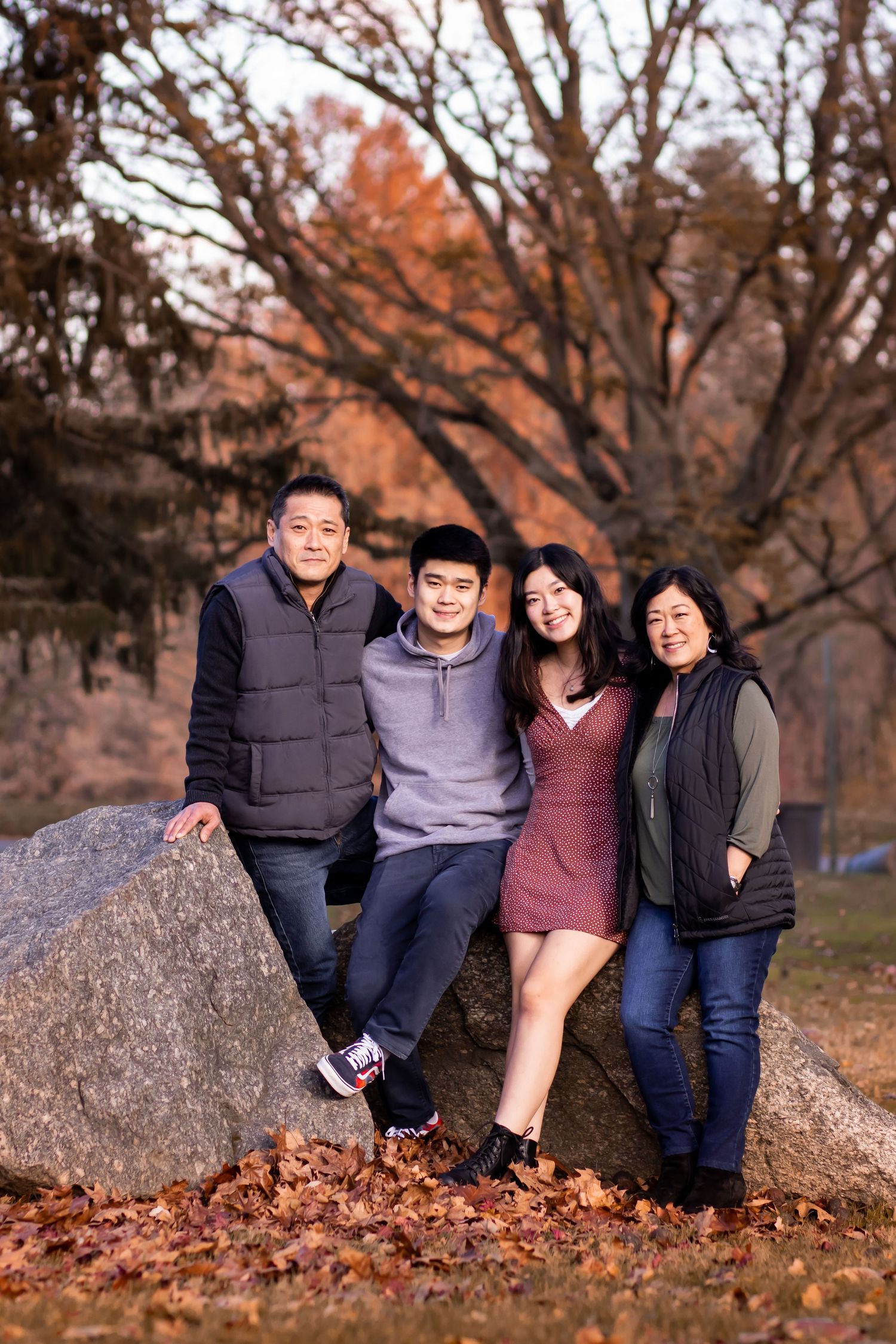 A family sits together on a large rock during autumn with fallen leaves scattered on the ground during a photo session.