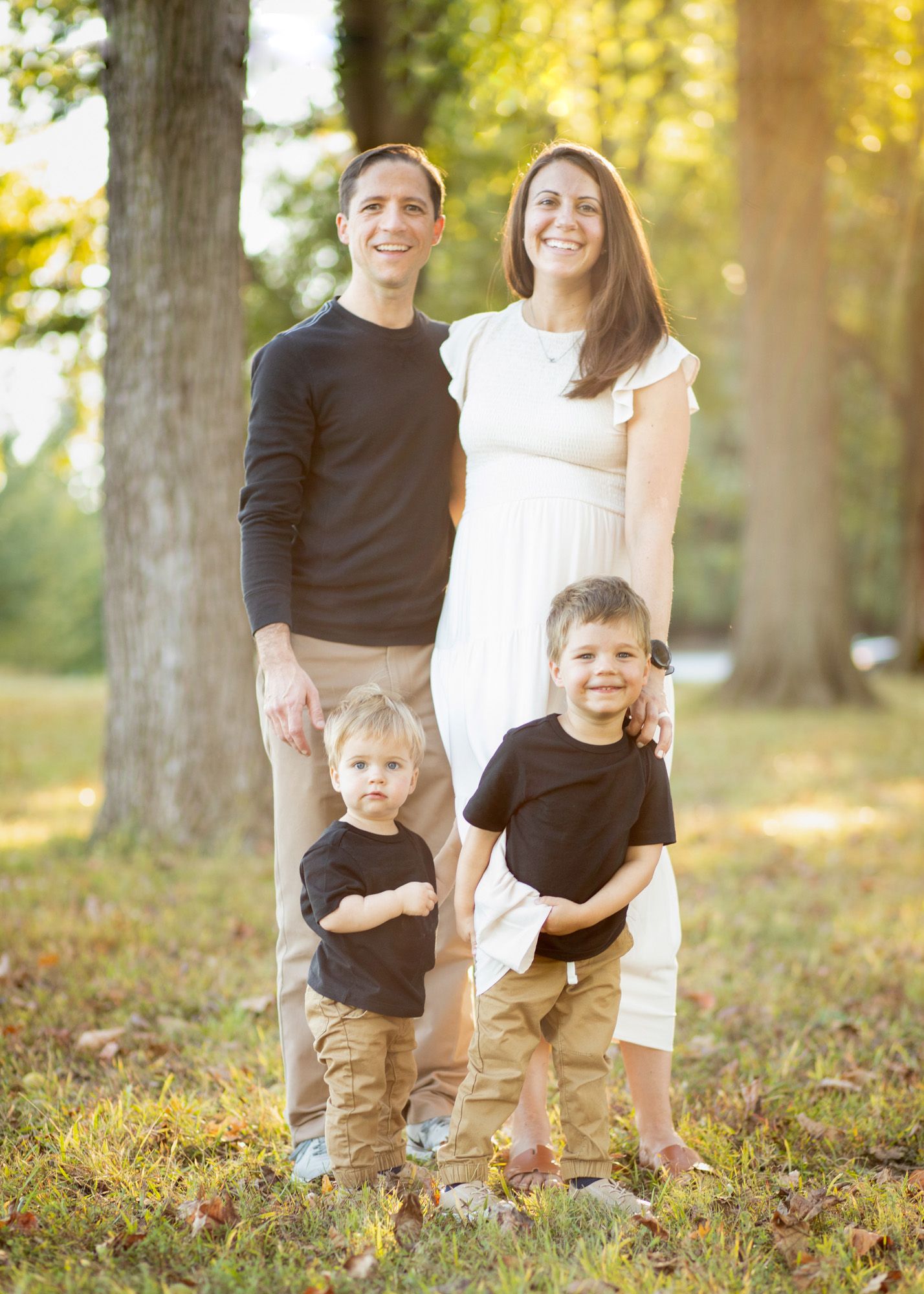 Family of four posing outdoors in golden hour light, parents with two young boys in matching outfits.