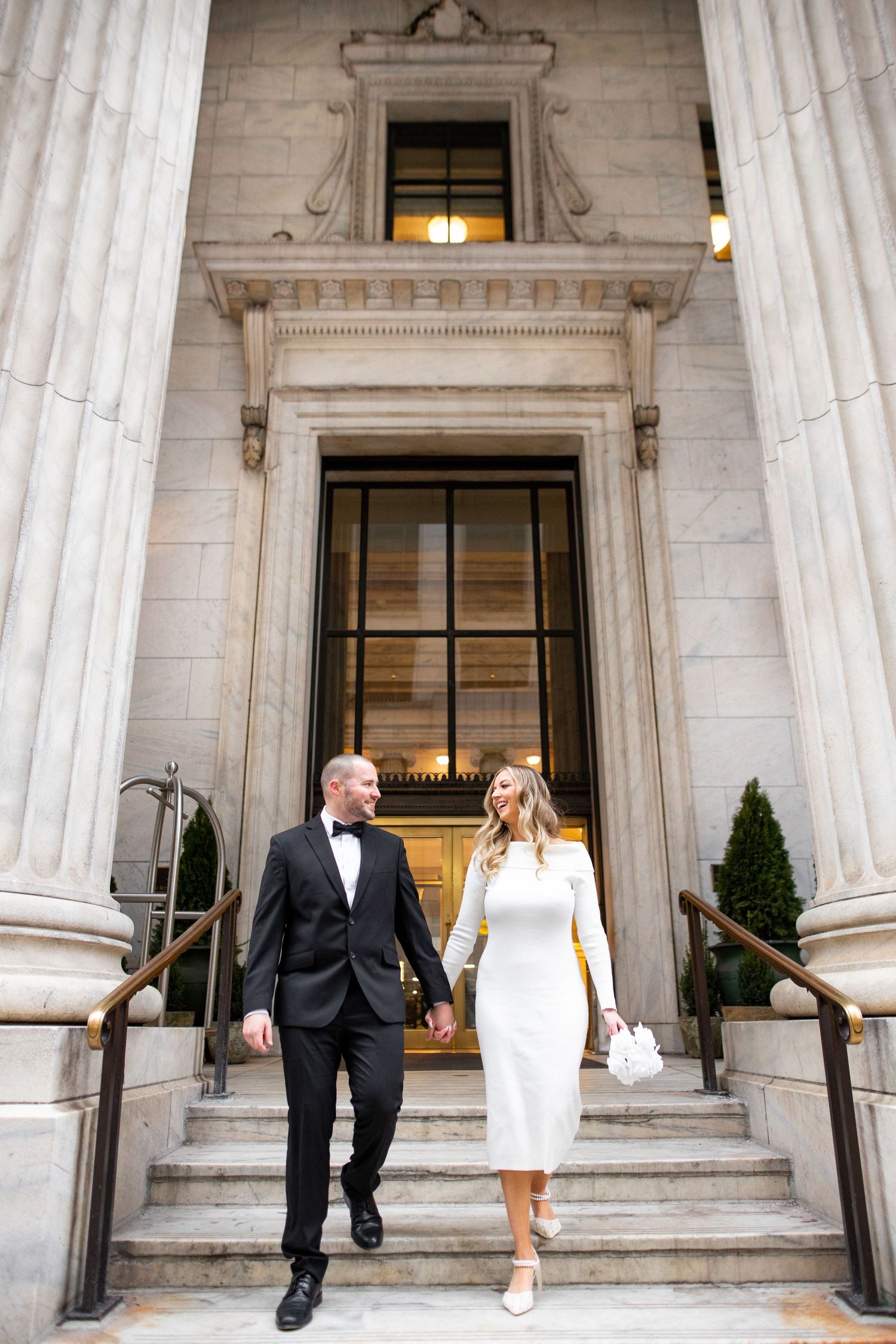 Elegant couple walking down historic building steps in formal attire with columns and grand entrance.