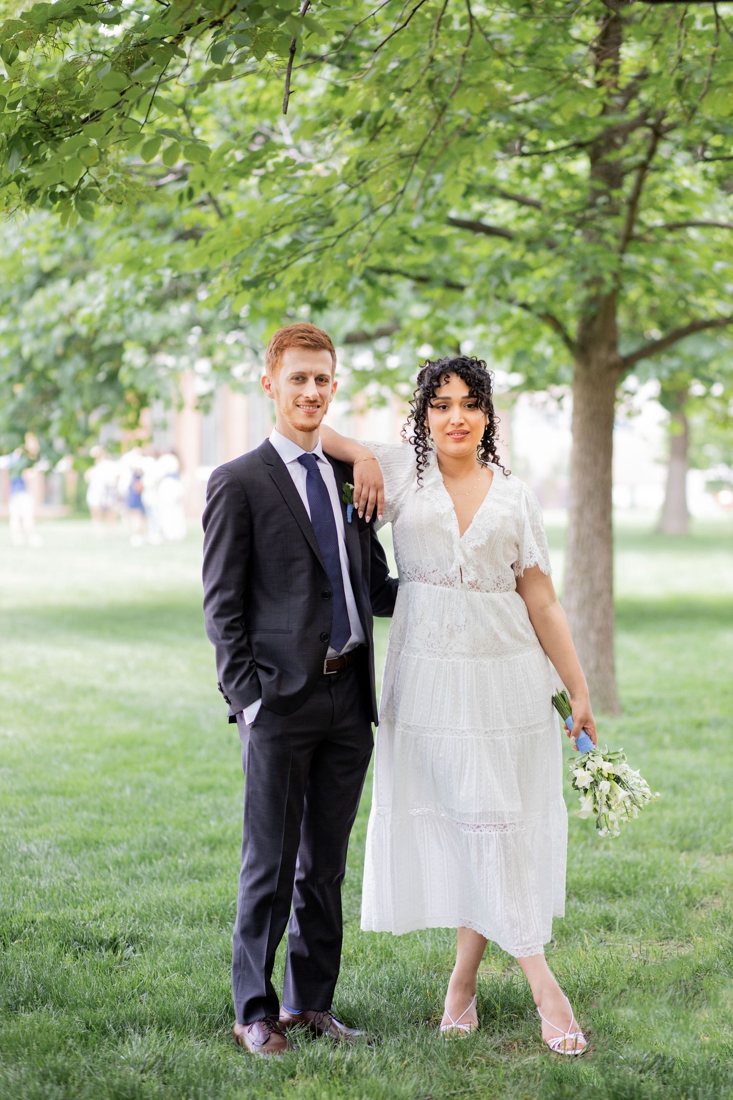 A couple in wedding attire poses for romantic shots under lush green trees in a park setting.