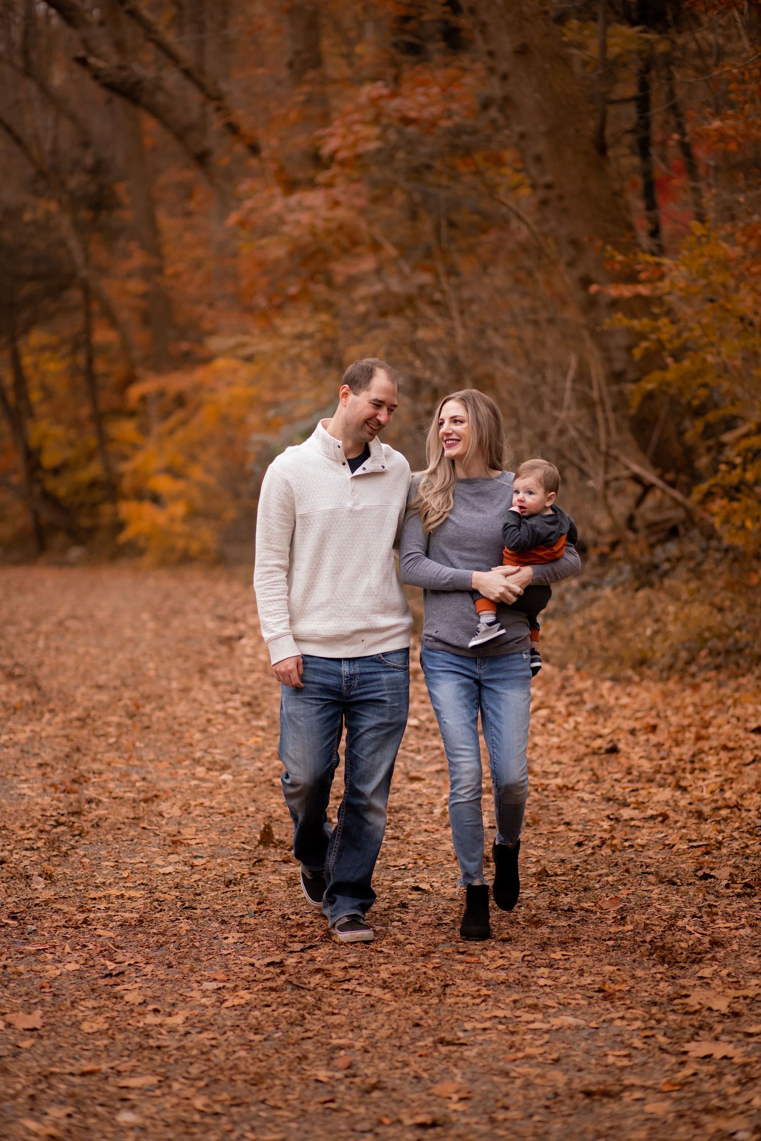 A couple walking together on a leaf-covered autumn trail in the woods while carrying a small child.