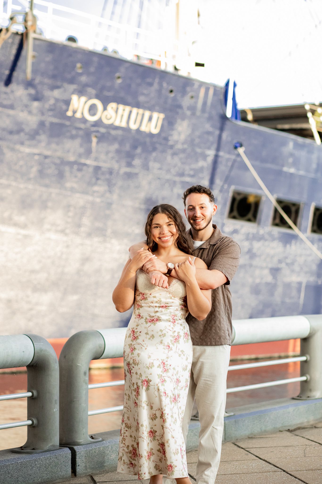 A couple poses playfully on a pier with a blue ship in the background.