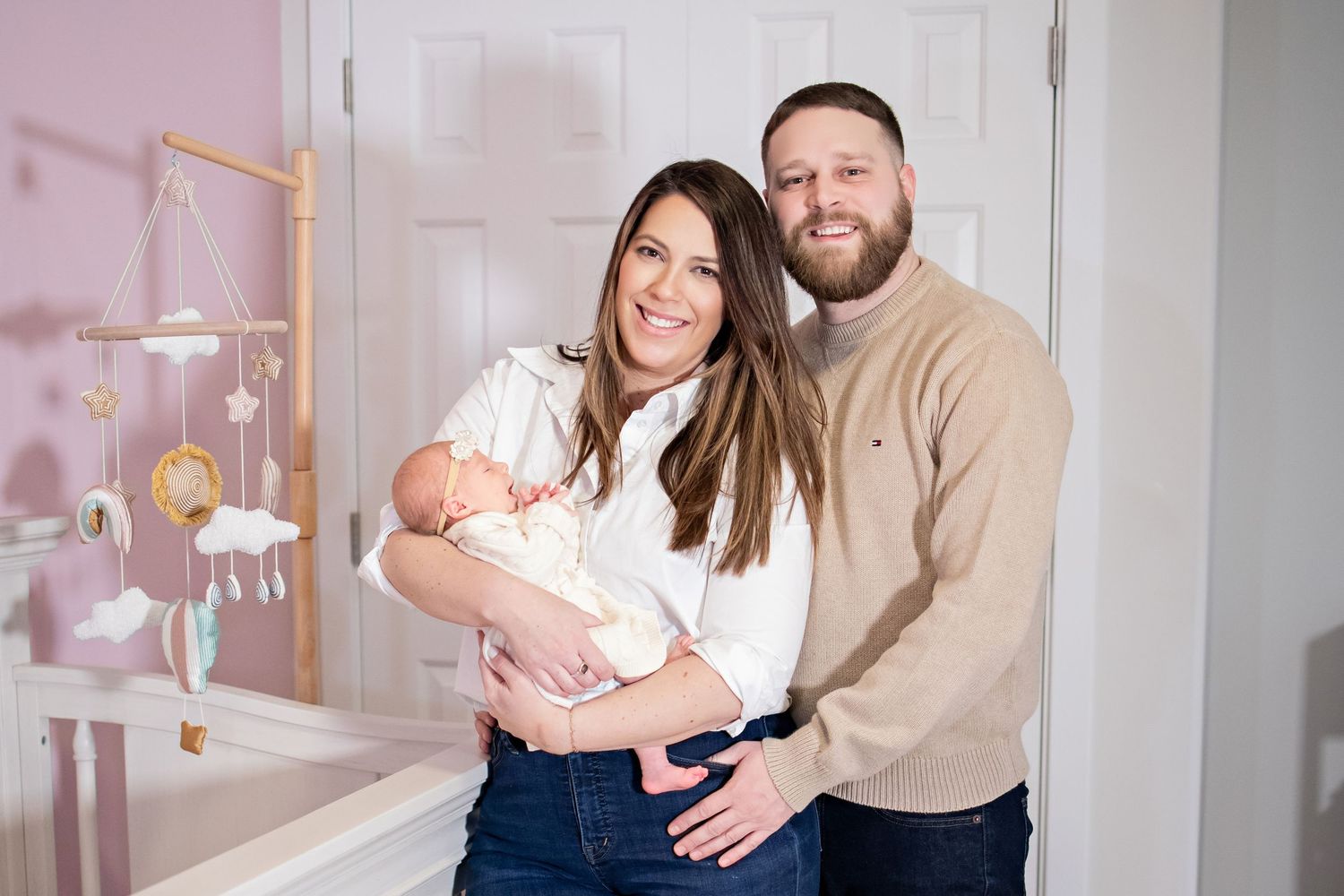 A loving couple in matching beige sweaters holds their newborn baby in a bright nursery setting.