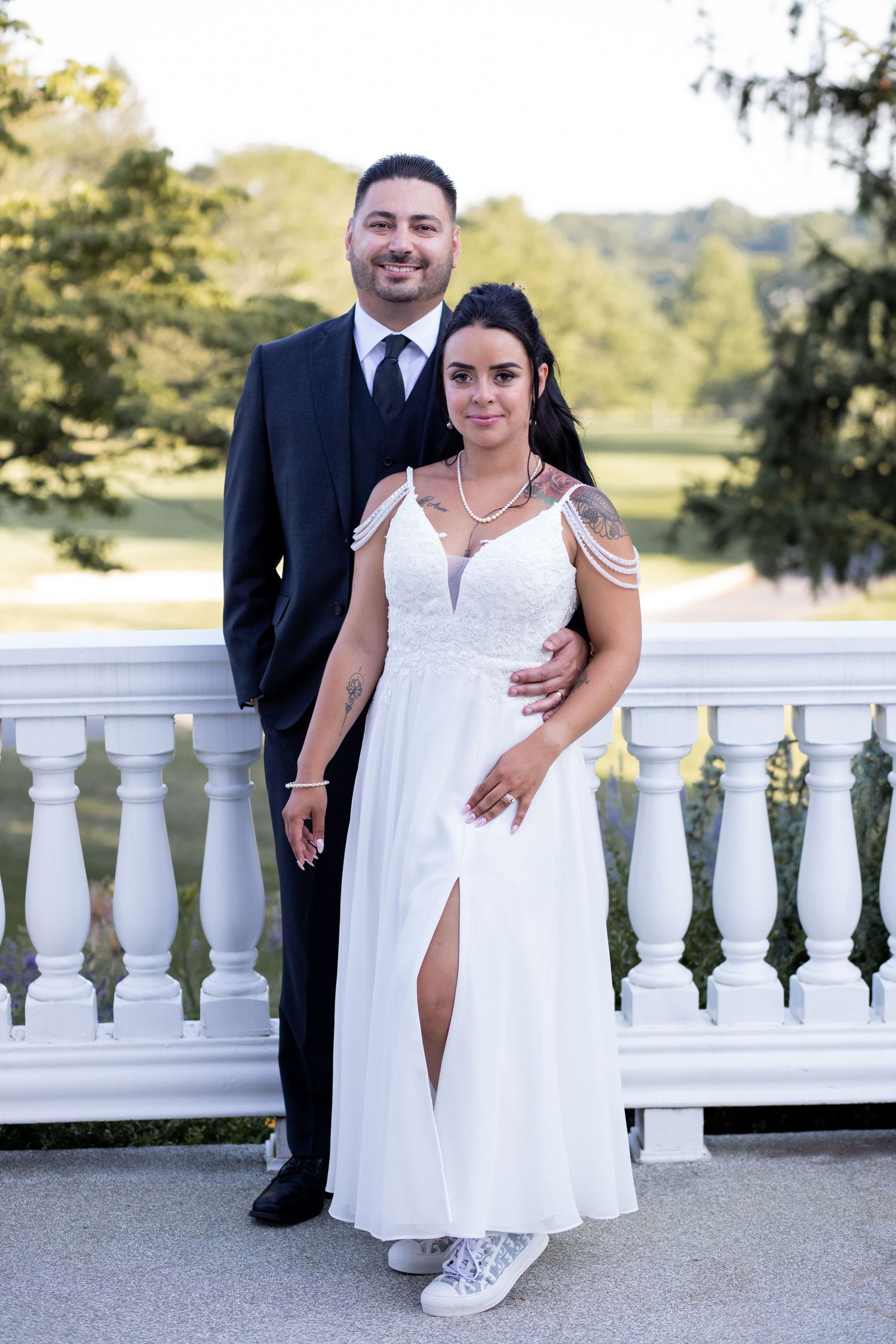 A couple poses outdoors on a white balcony railing, with the bride in a white dress and groom in a dark suit.