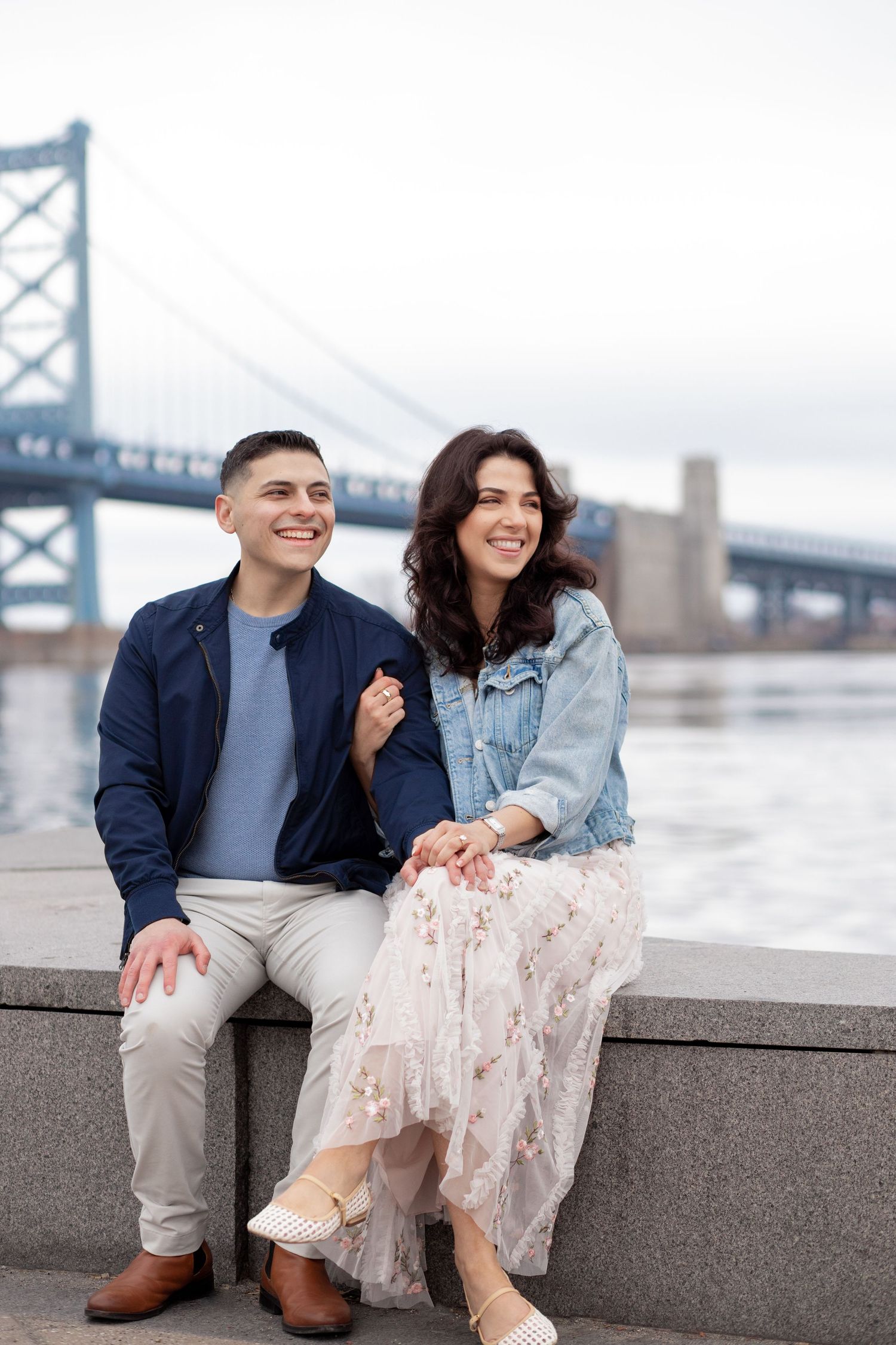A couple sits together near a bridge, smiling and enjoying a cloudy day by the waterfront.