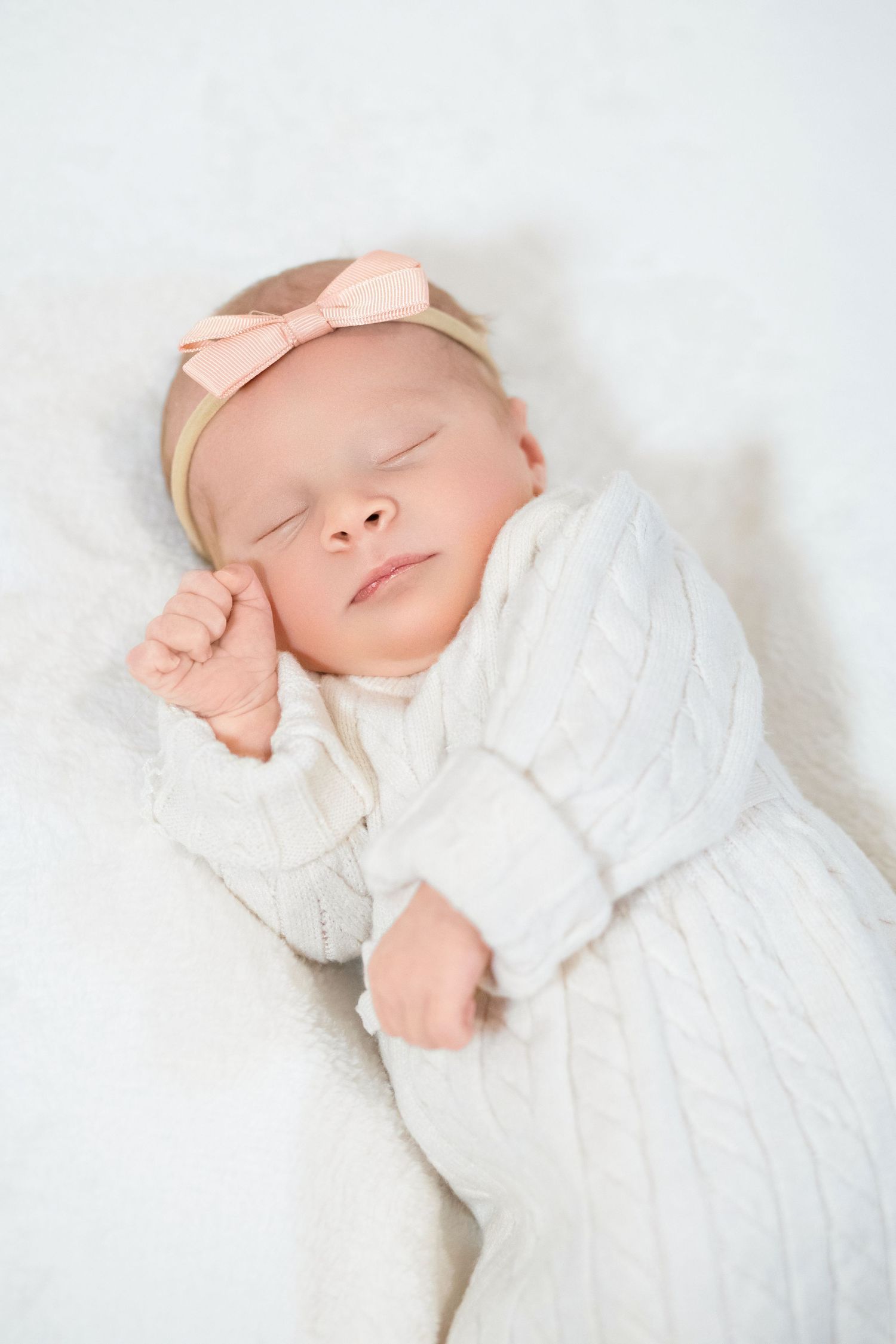 Sleeping newborn baby wearing white knit sweater and peach bow headband rests peacefully on white blanket.