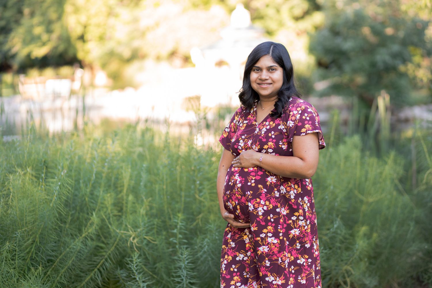 Pregnant woman in floral dress cradling baby bump outdoors in lush green garden during maternity photo session.