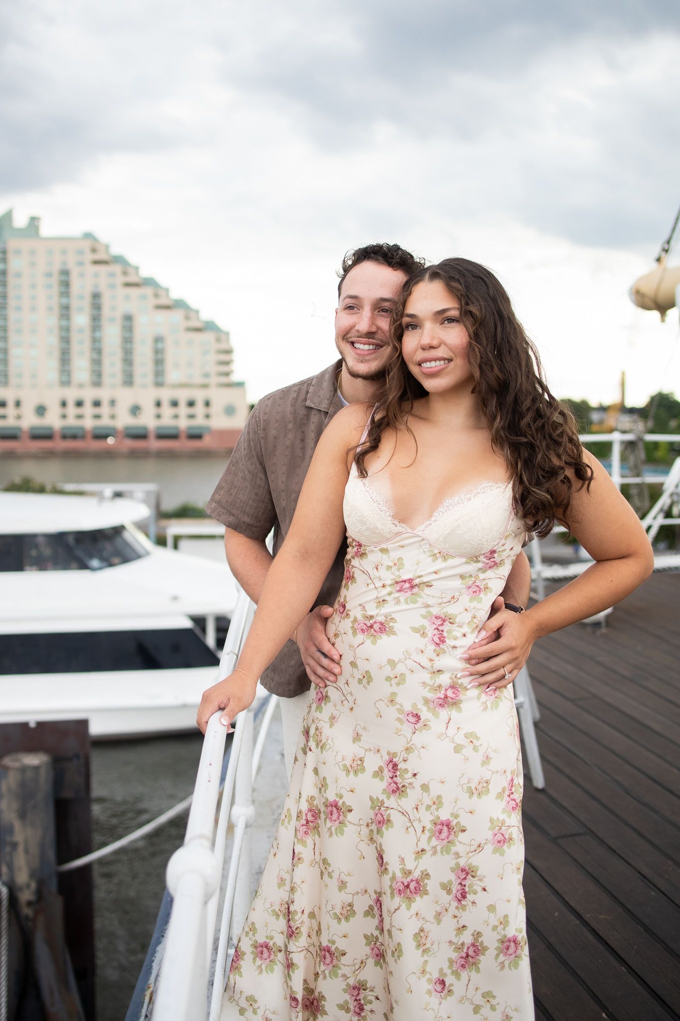A couple poses together on a wooden dock with boats and city buildings visible in the background on a cloudy day.