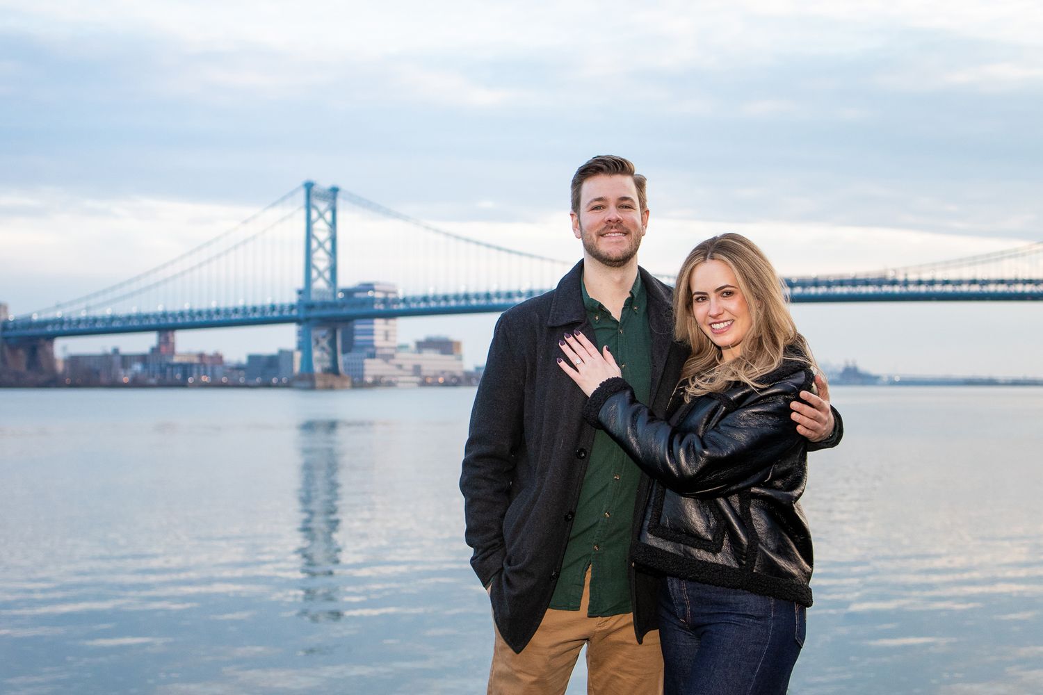 A couple poses together near a waterfront with a large suspension bridge visible in the background.