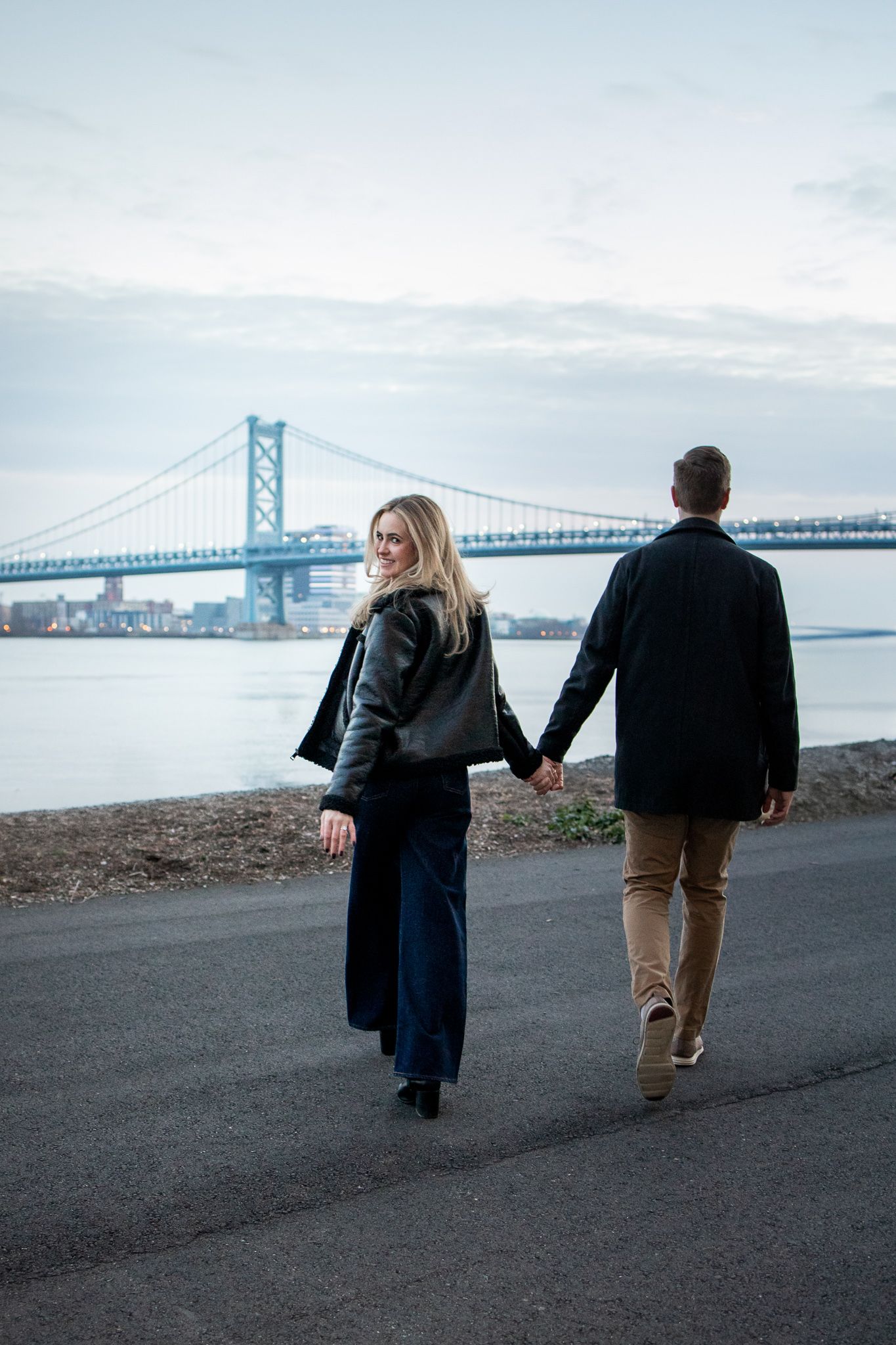 Two people walking along a waterfront path with the Bay Bridge spanning across the horizon.