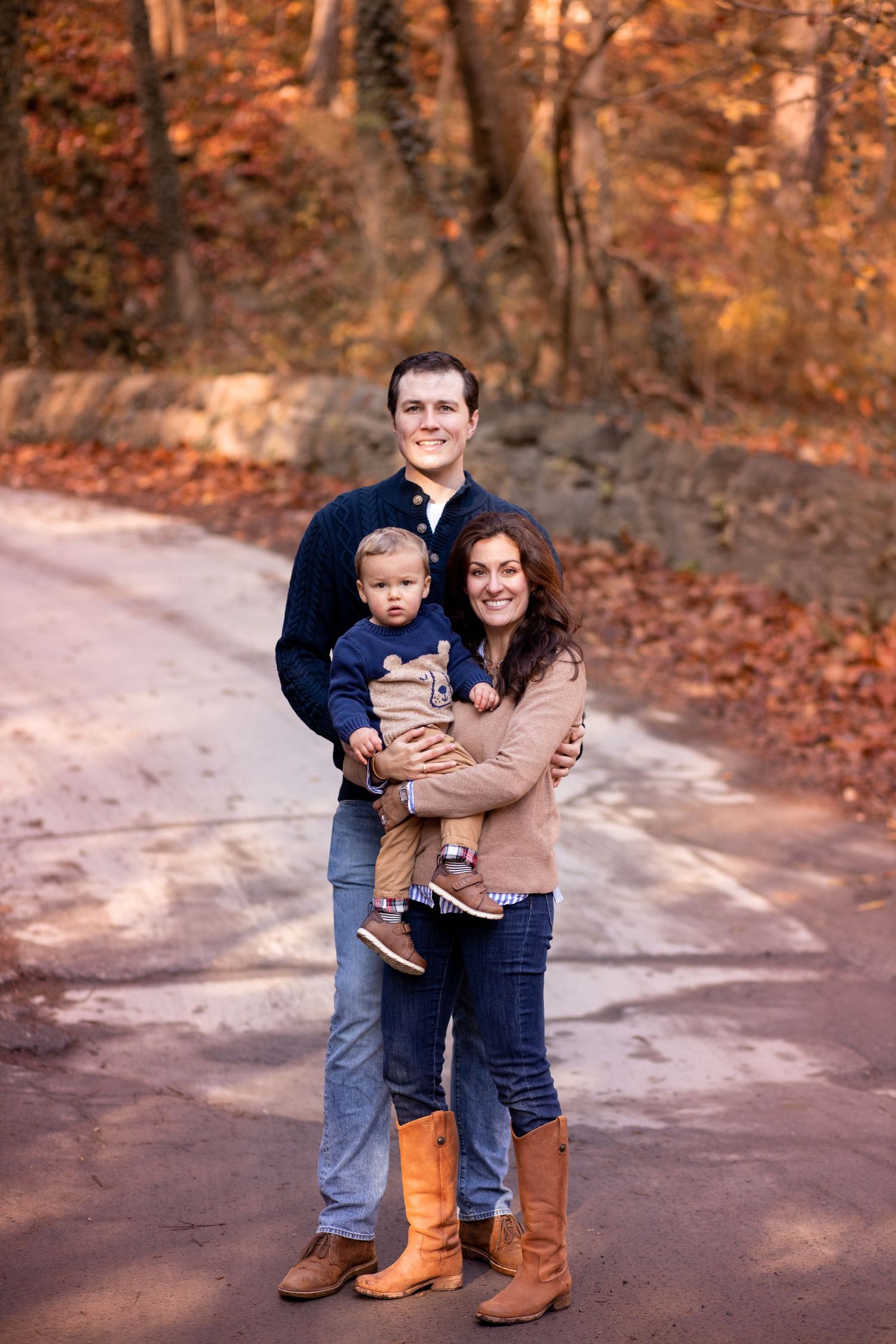 A family stands together on a wooded path during autumn with orange fallen leaves in the background.