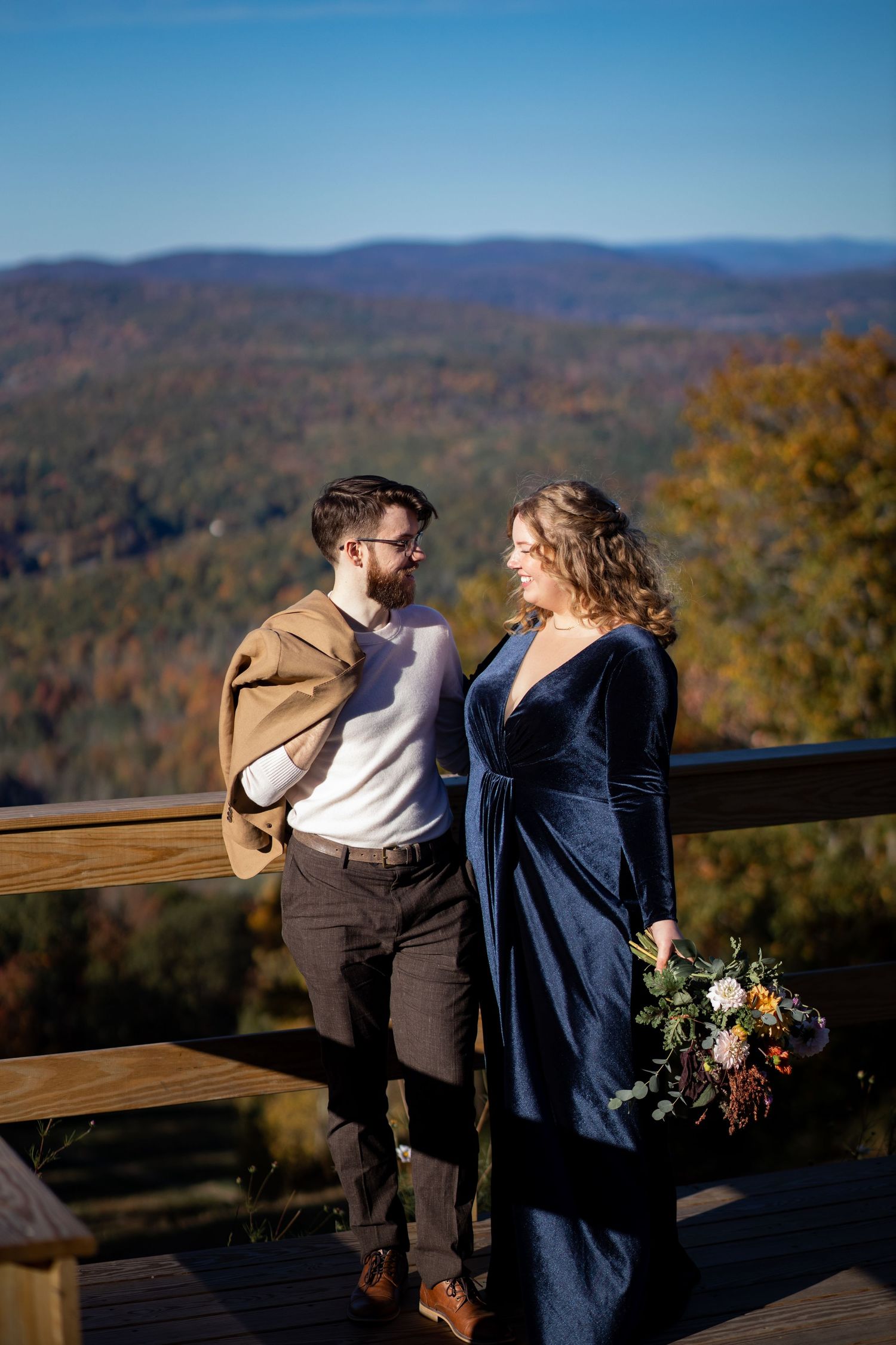 A couple stands on a wooden deck overlooking autumn mountain vistas with fall foliage in the background.