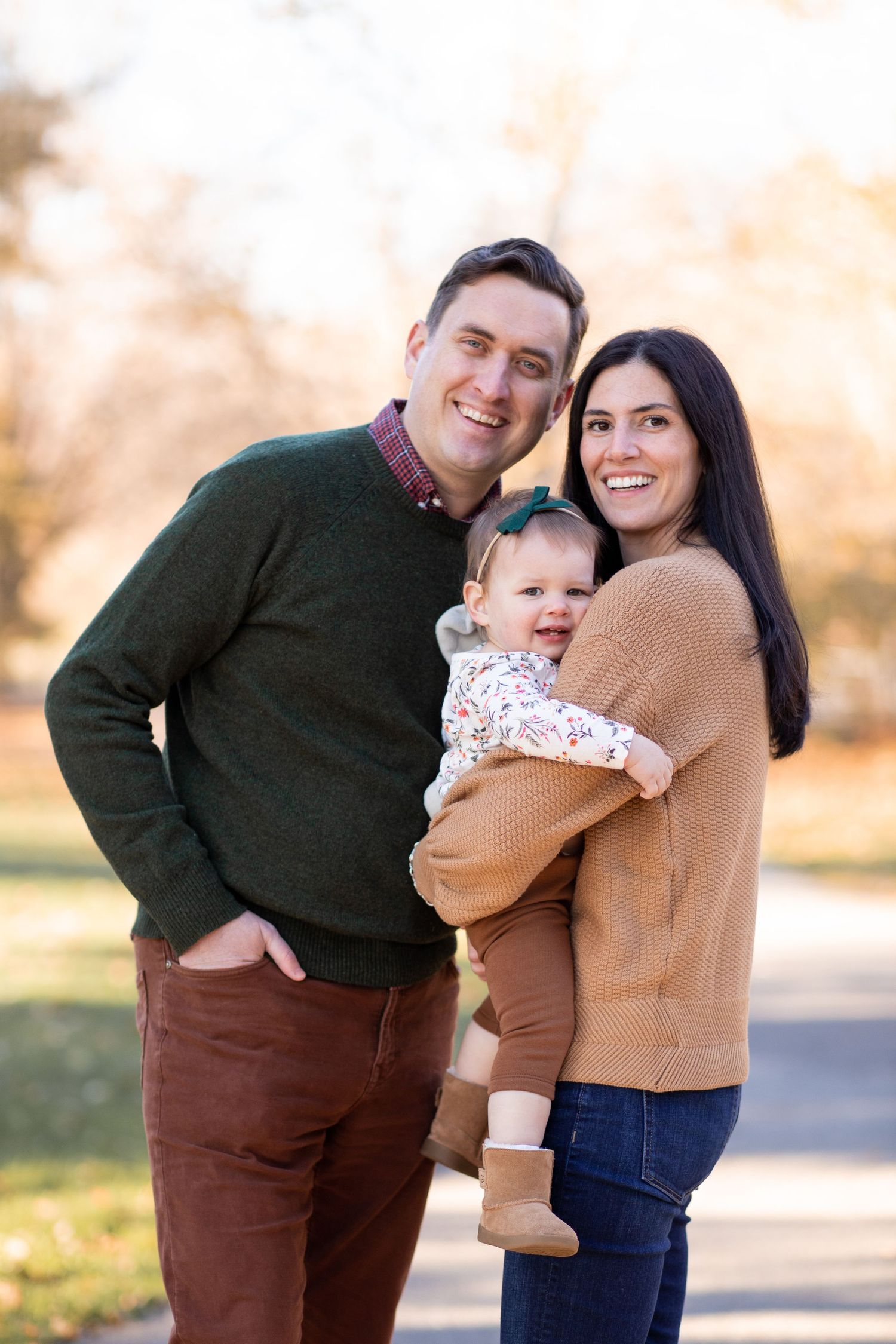 A family wearing coordinating sweaters poses together outdoors on a sunny autumn day in a park.
