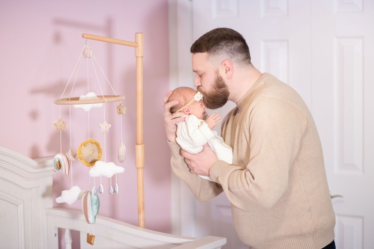 A person wearing beige adjusts a pink and gold mobile hanging above a white crib in a nursery.