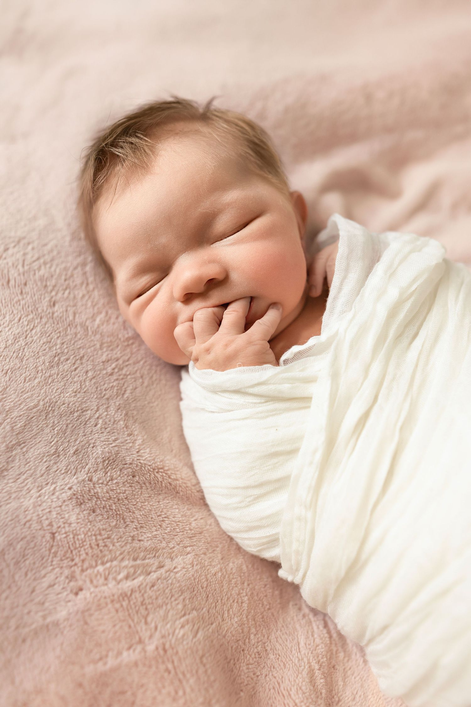Peaceful sleeping newborn wrapped in soft white swaddle blanket resting on pink textured surface.